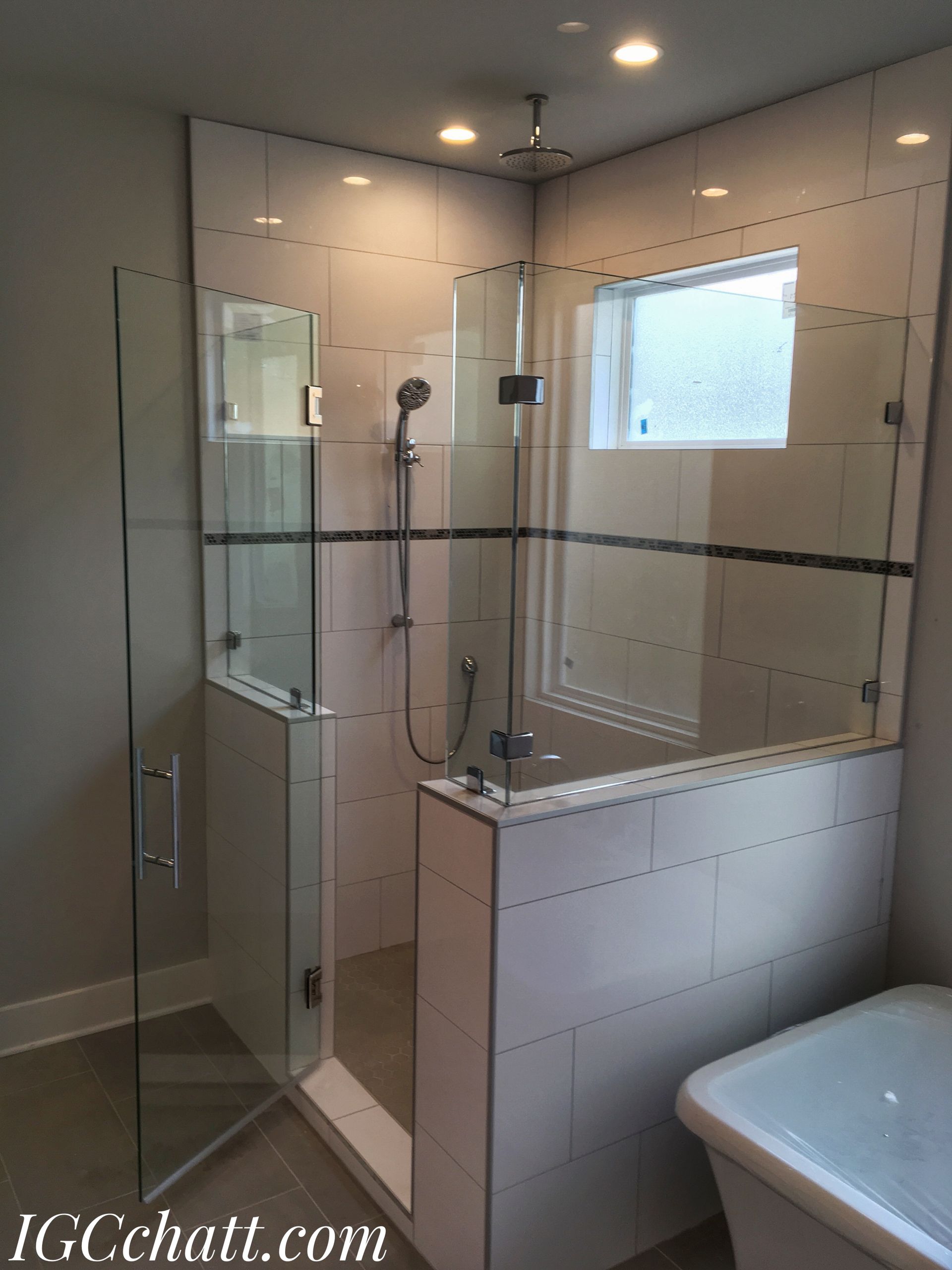 A modern bathroom with a glass-enclosed shower. Beige tiled walls and a small window are visible.