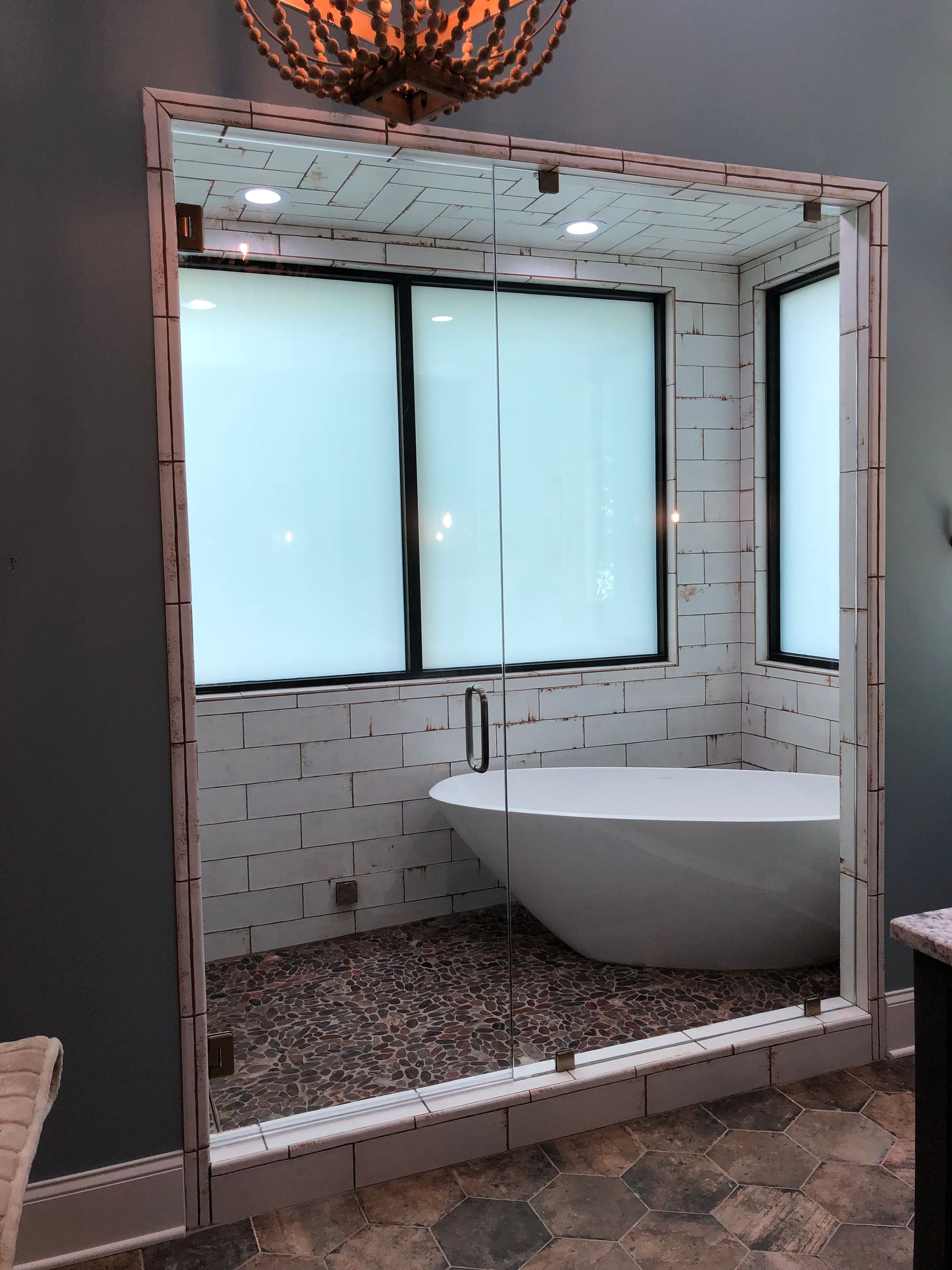 A modern bathroom with a glass-enclosed shower/bath area. White soaking tub, pebble floor, and tiled walls are visible.