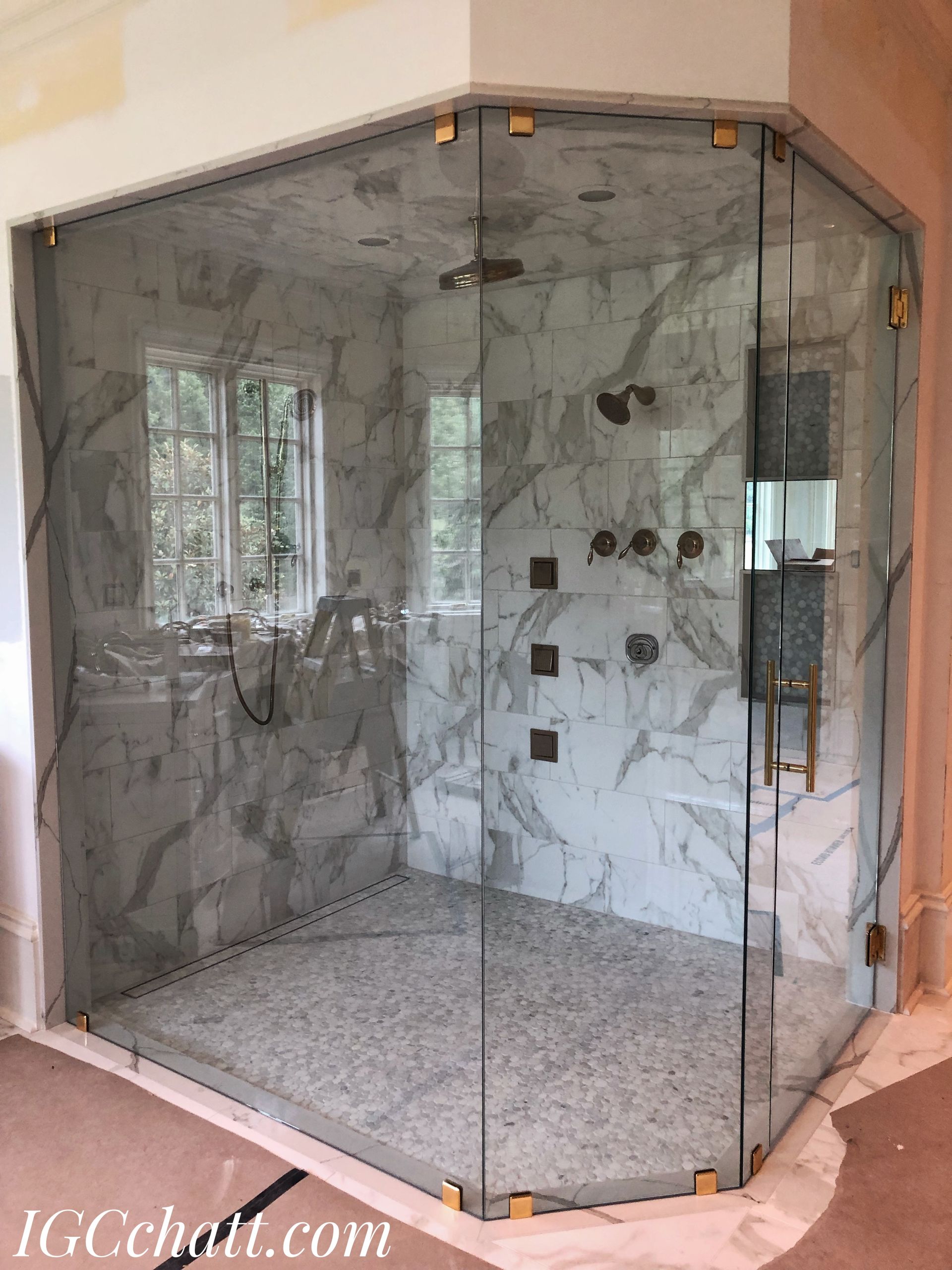 Corner shower with marble walls and glass doors, gold hardware. A window is visible behind the glass.