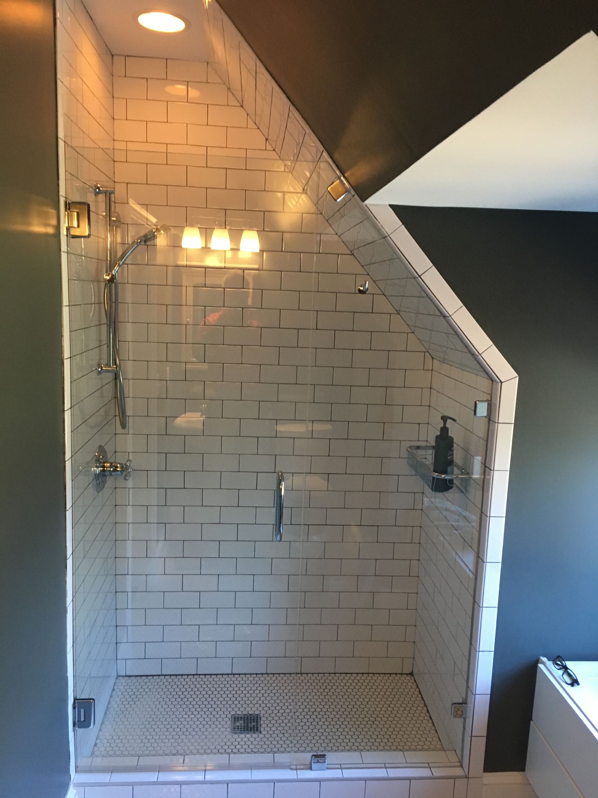 Tiled shower with a glass door. White subway tile covers the walls, which slope inward at the top.