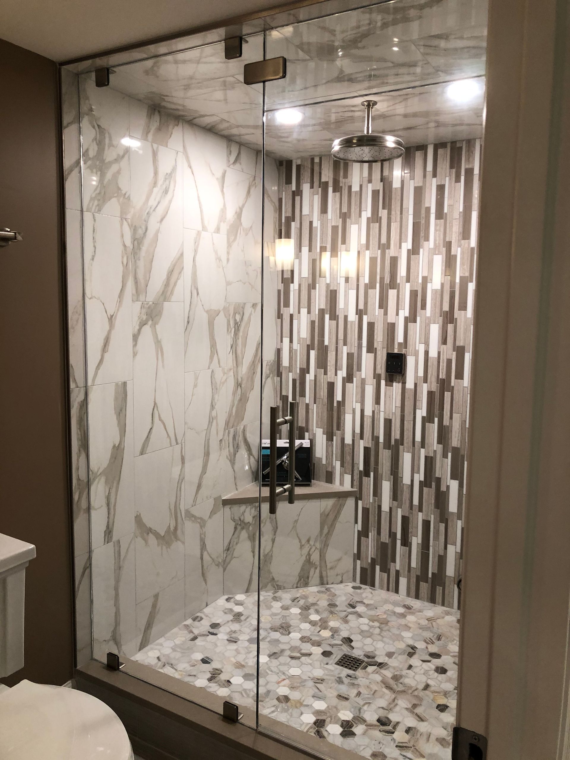 A modern shower with a glass door. Features marble-patterned tile and a pebbled floor, with a mosaic tile accent wall.