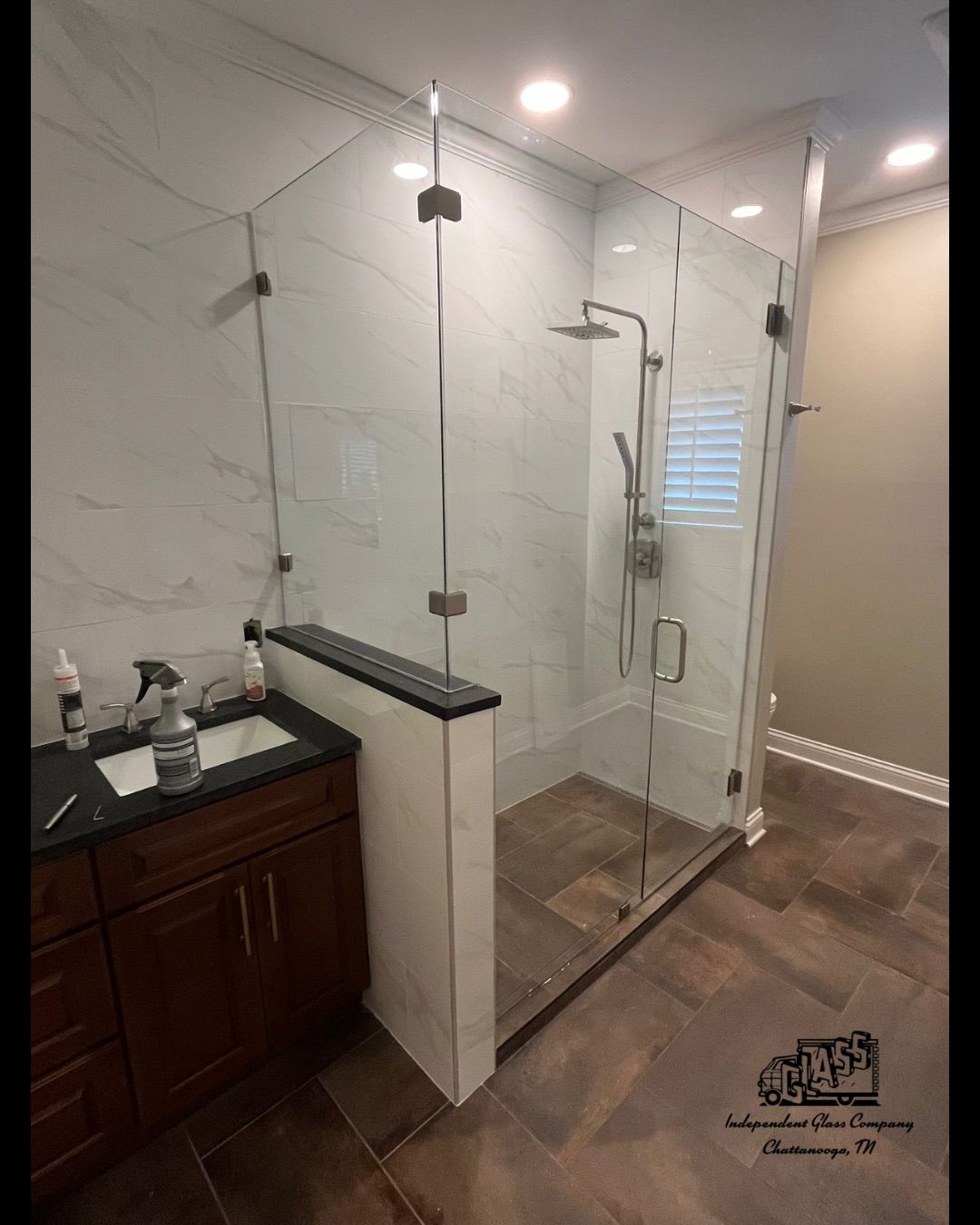 A modern bathroom with a glass shower enclosure, brown floor tiles, and marble-like wall tiles. A dark wood vanity sits to the left.