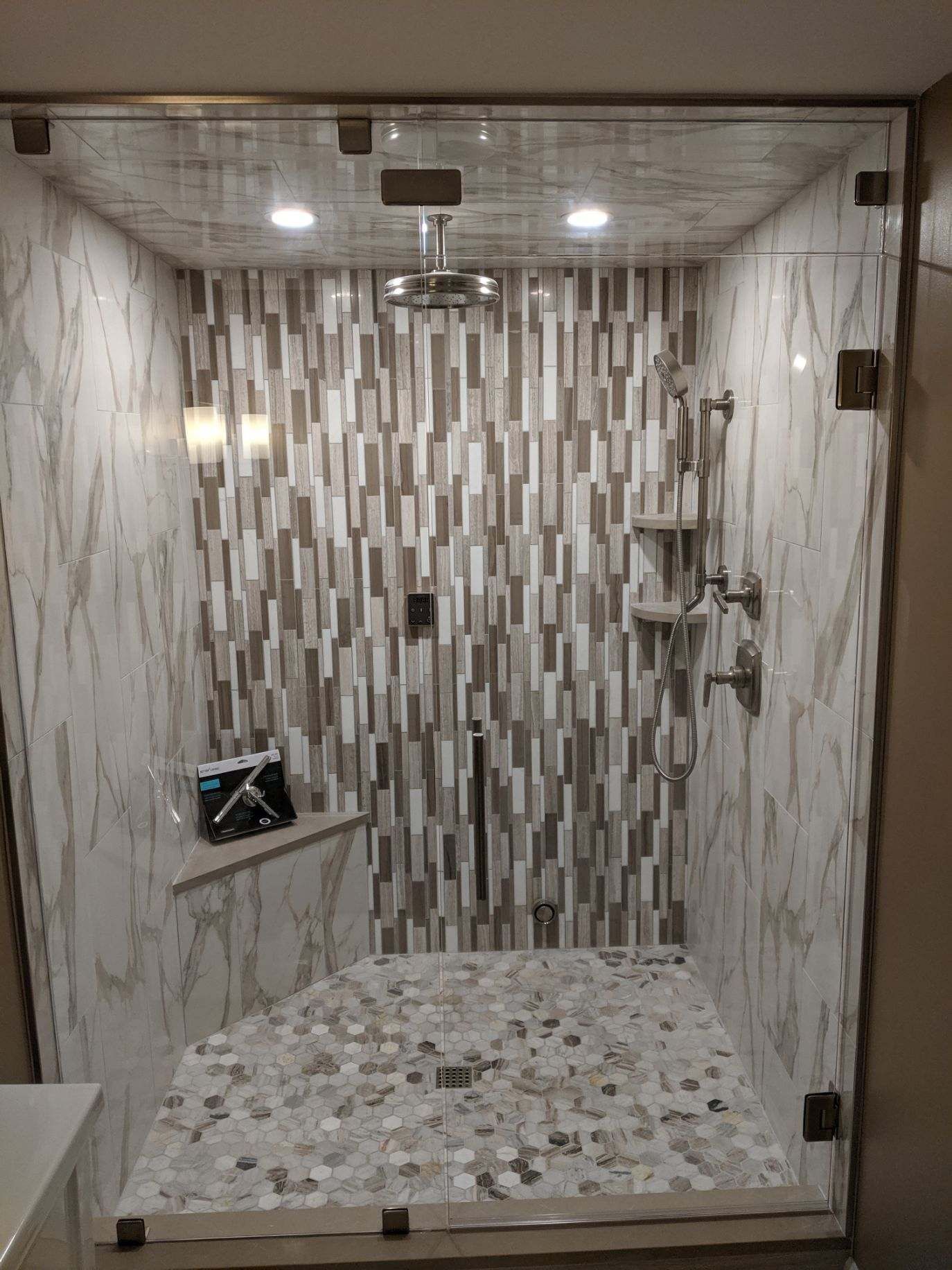A modern shower with glass doors, featuring mosaic and marble-look tiles, and a pebble-tiled floor.