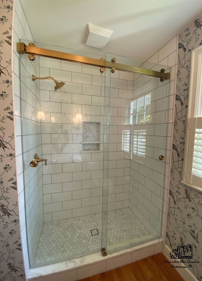 Shower stall with glass doors and gold hardware; subway tile walls. Small window to the right; floral wallpaper.