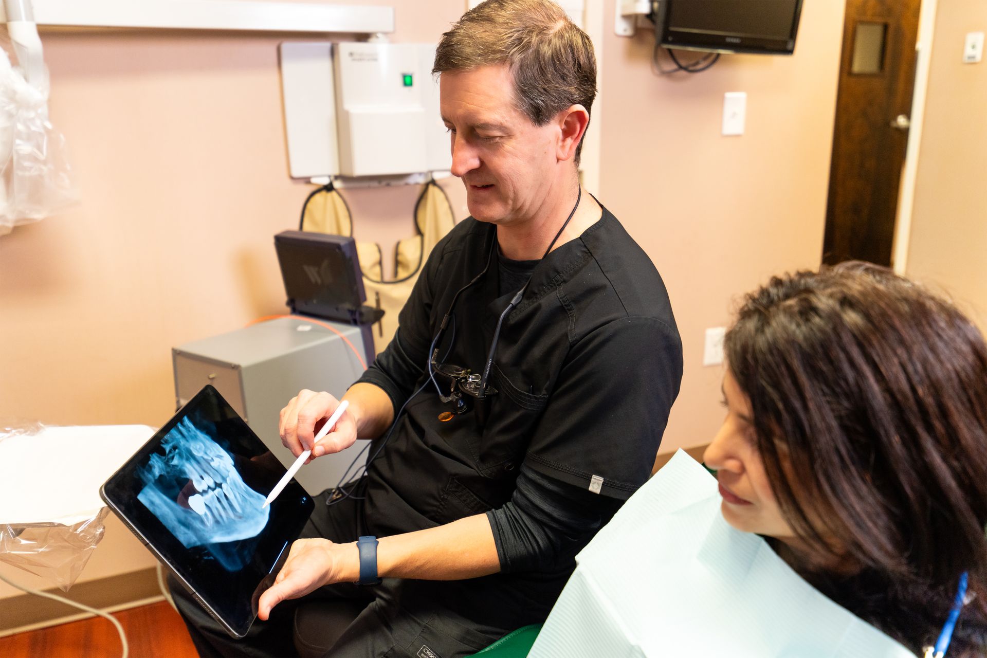 A dental professional uses a stylus to point to a digital X-ray on a tablet while consulting with a patient in an office.