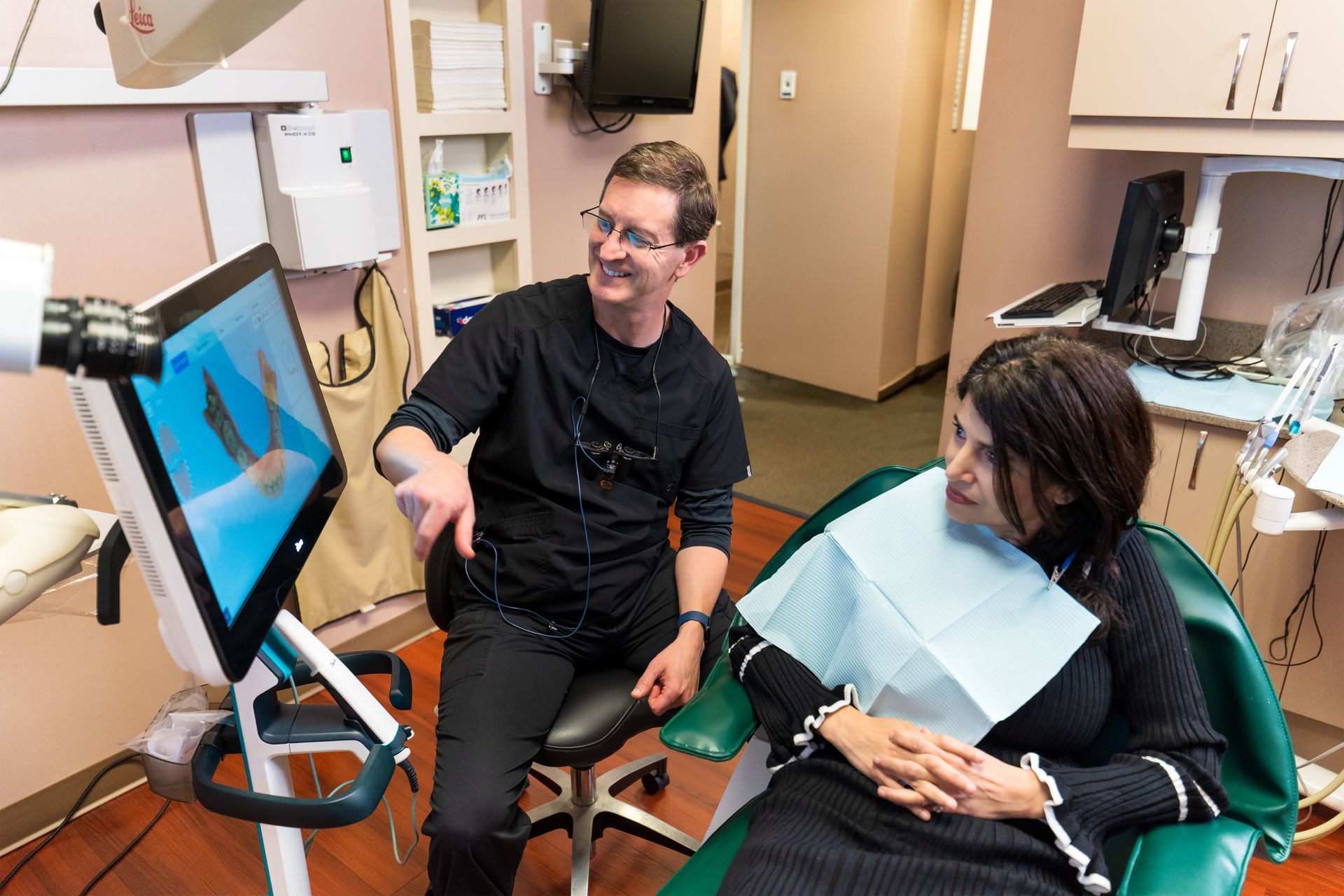 A dentist points to a 3D dental scan on a screen while consulting with a patient in an office.