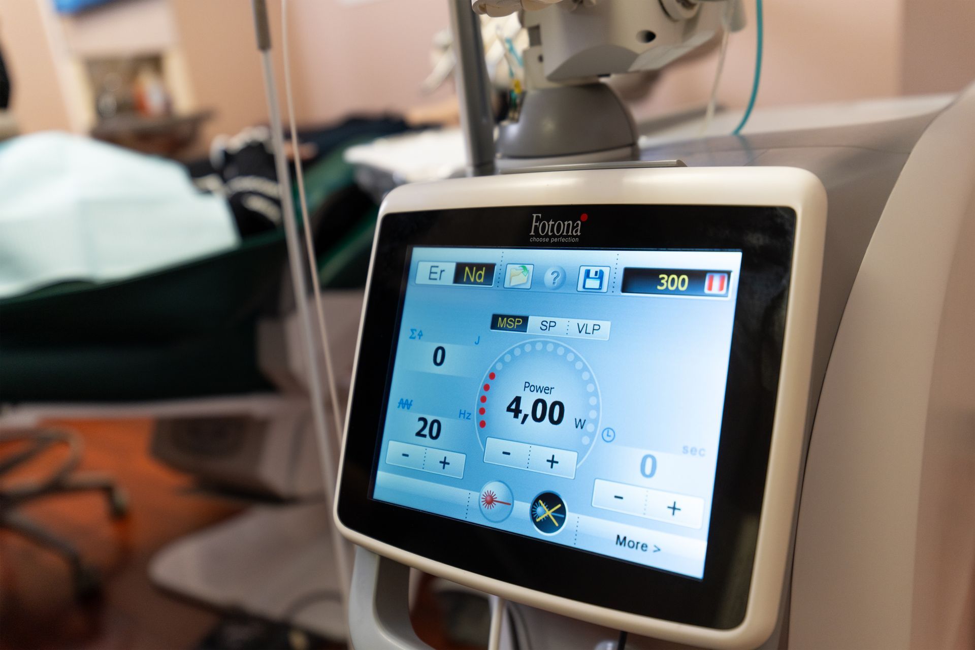 A close-up of a medical laser device's screen showing settings, with a blurry clinical treatment room in the background.