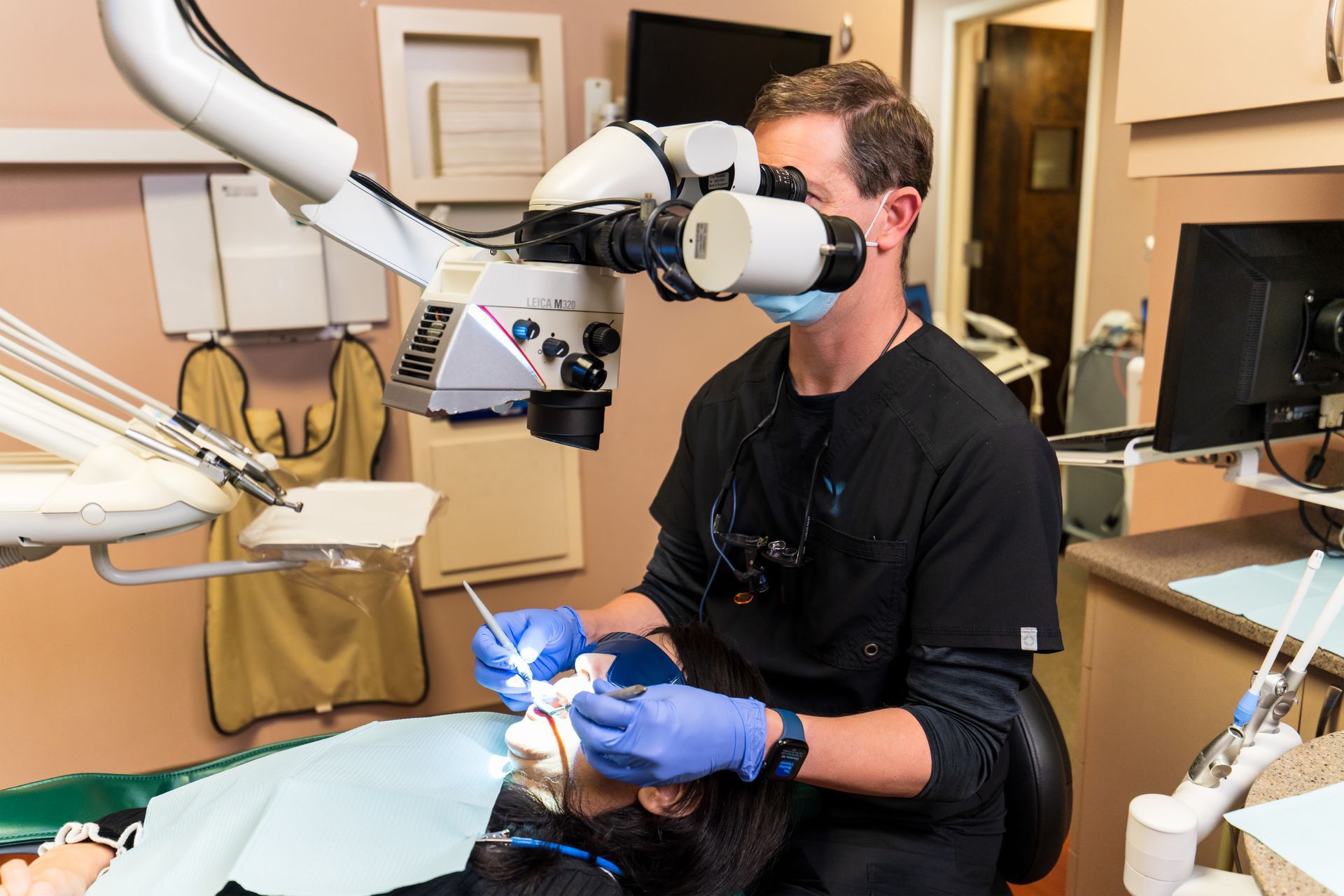 A dentist in blue gloves uses a dental microscope to perform a procedure on a patient in a clinical exam room.