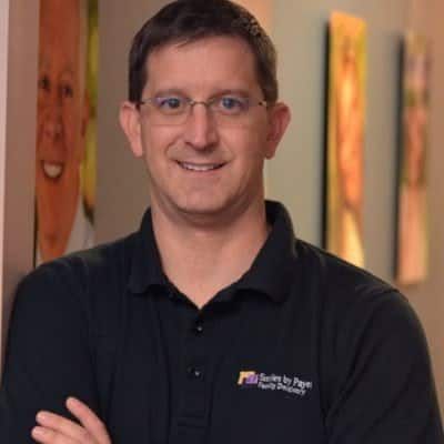 Man with glasses and black shirt smiles, arms crossed;