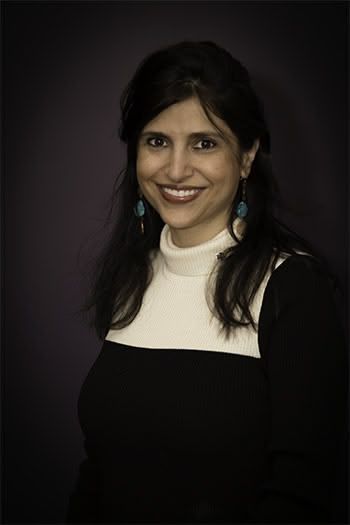 Woman smiling, wearing a black and white sweater, turquoise earrings, against a purple backdrop.