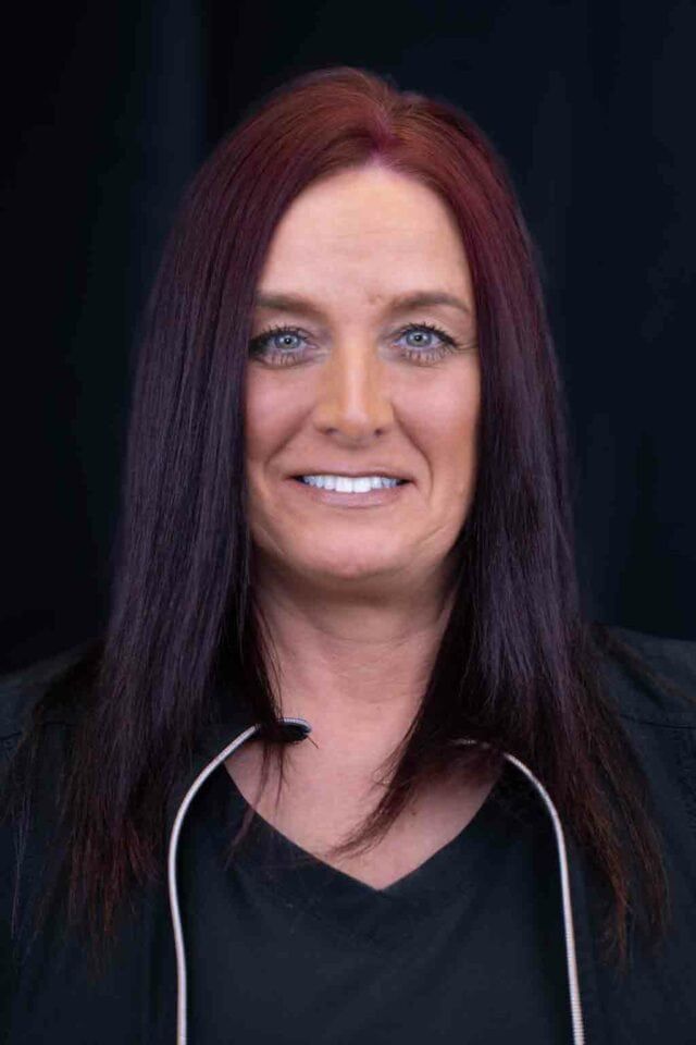 Woman with long, reddish-purple hair smiles, wearing a black top. A white cord hangs around her neck against a black backdrop.