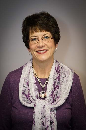 Woman with short, dark hair, glasses, and a purple scarf smiles.