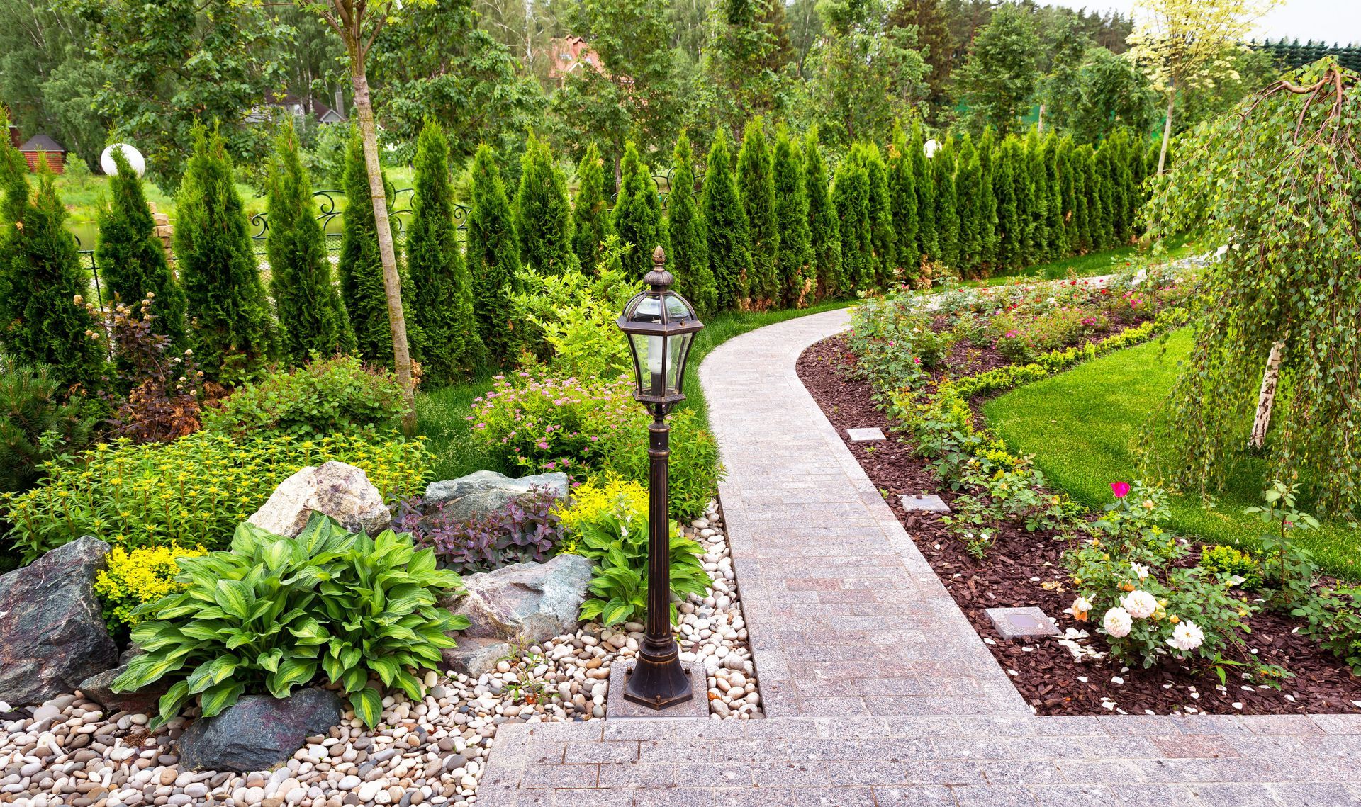A stone pathway curves through a lush garden with a central lamp post, manicured hedges, and decorative rocks.