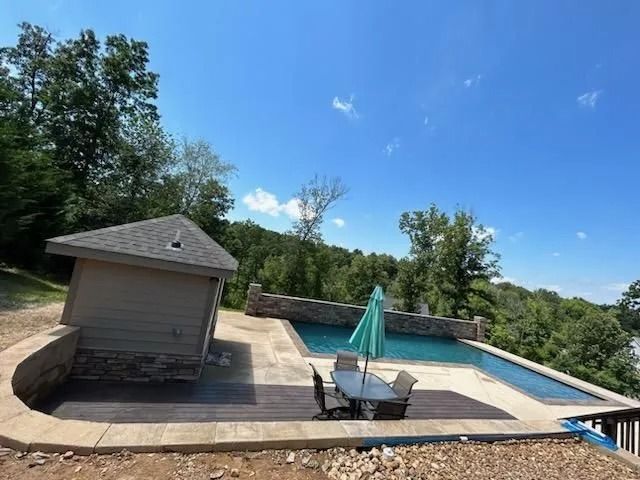 A swimming pool and patio area with a tan pool house, stone retaining wall, and a patio set with a teal umbrella.