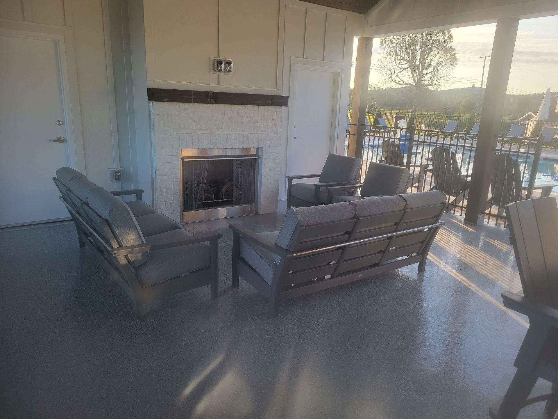 A covered patio with a fireplace and grey outdoor furniture set, overlooking an outdoor pool area at sunset.