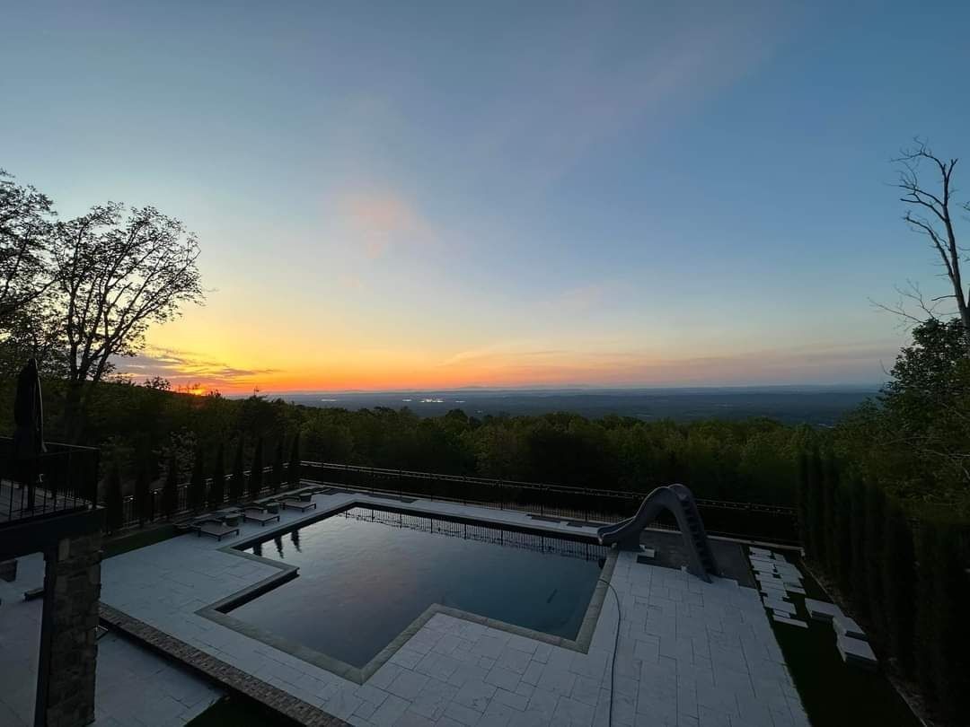 A rectangular swimming pool sits on a patio overlooking a lush, forested valley during a vibrant sunset.