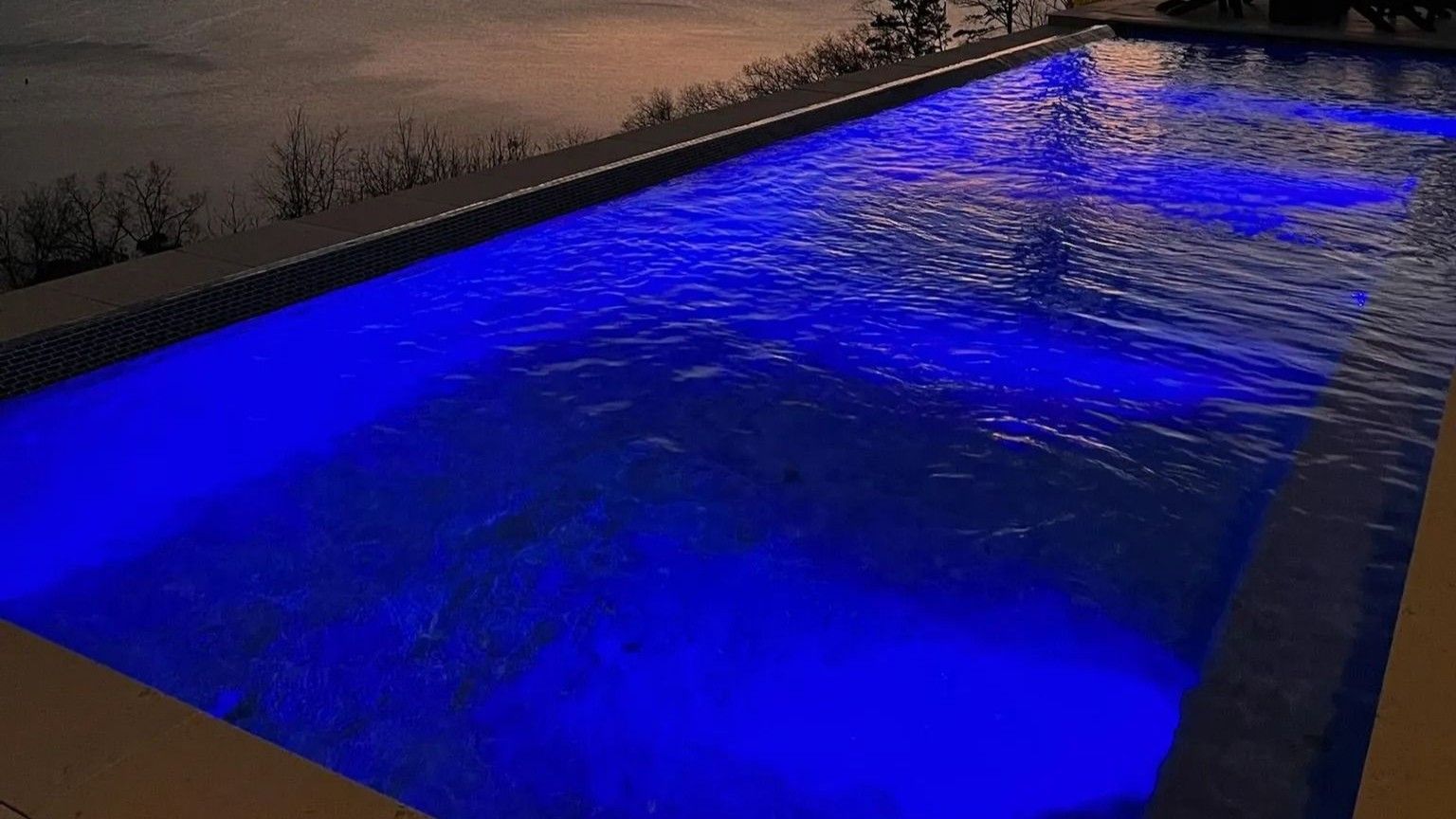 A rectangular swimming pool at dusk, illuminated with vibrant blue underwater lights.