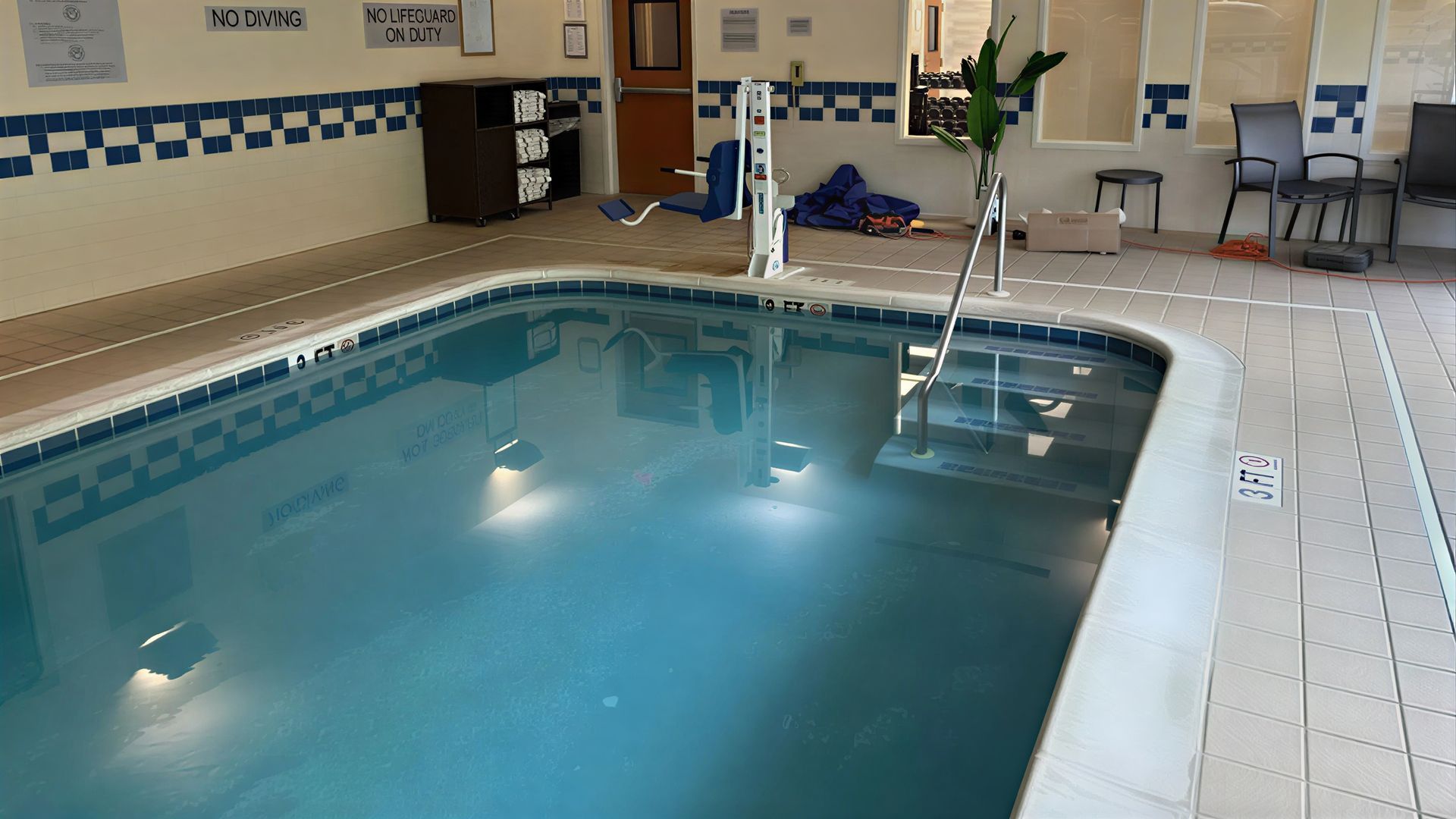 An indoor hotel swimming pool with blue water, a metal pool lift, and chairs on the surrounding tiled deck.