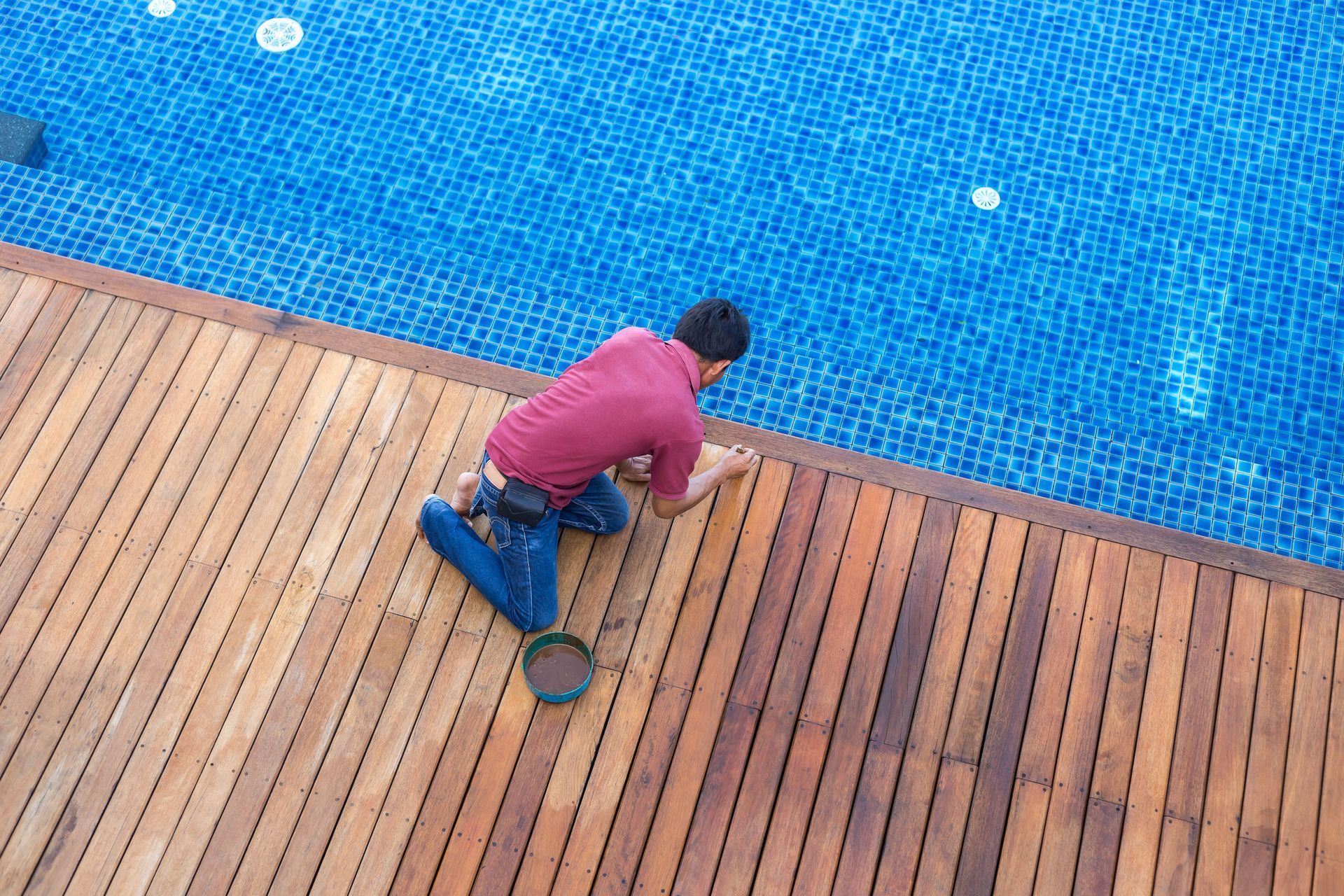 A person in a red shirt and blue jeans kneels on a wooden deck next to a blue swimming pool, applying oil to the wood.