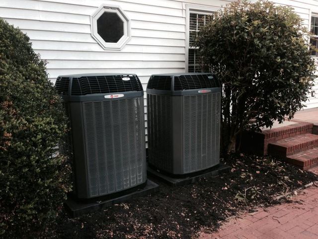 Two dark HVAC units on a brick foundation next to a white house with landscaping.