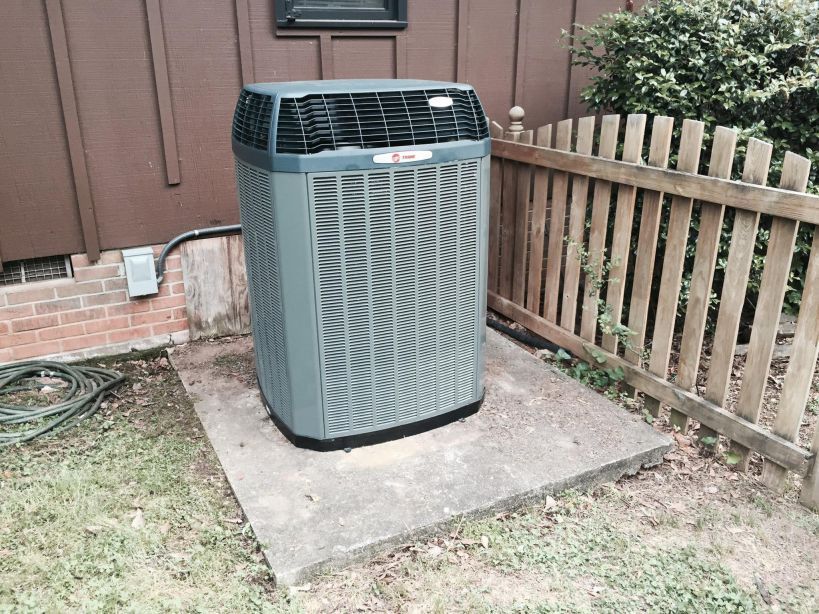Gray air conditioning unit on a concrete pad next to a wooden fence and a house.