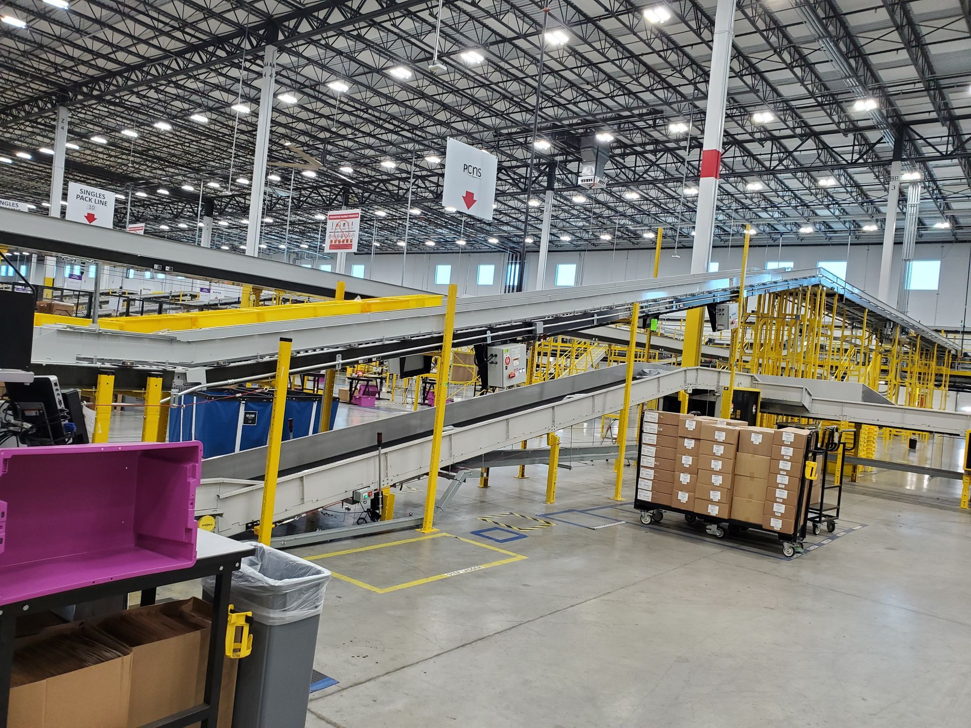 Large indoor warehouse with conveyor belts, yellow railings, and a pallet of cardboard boxes.