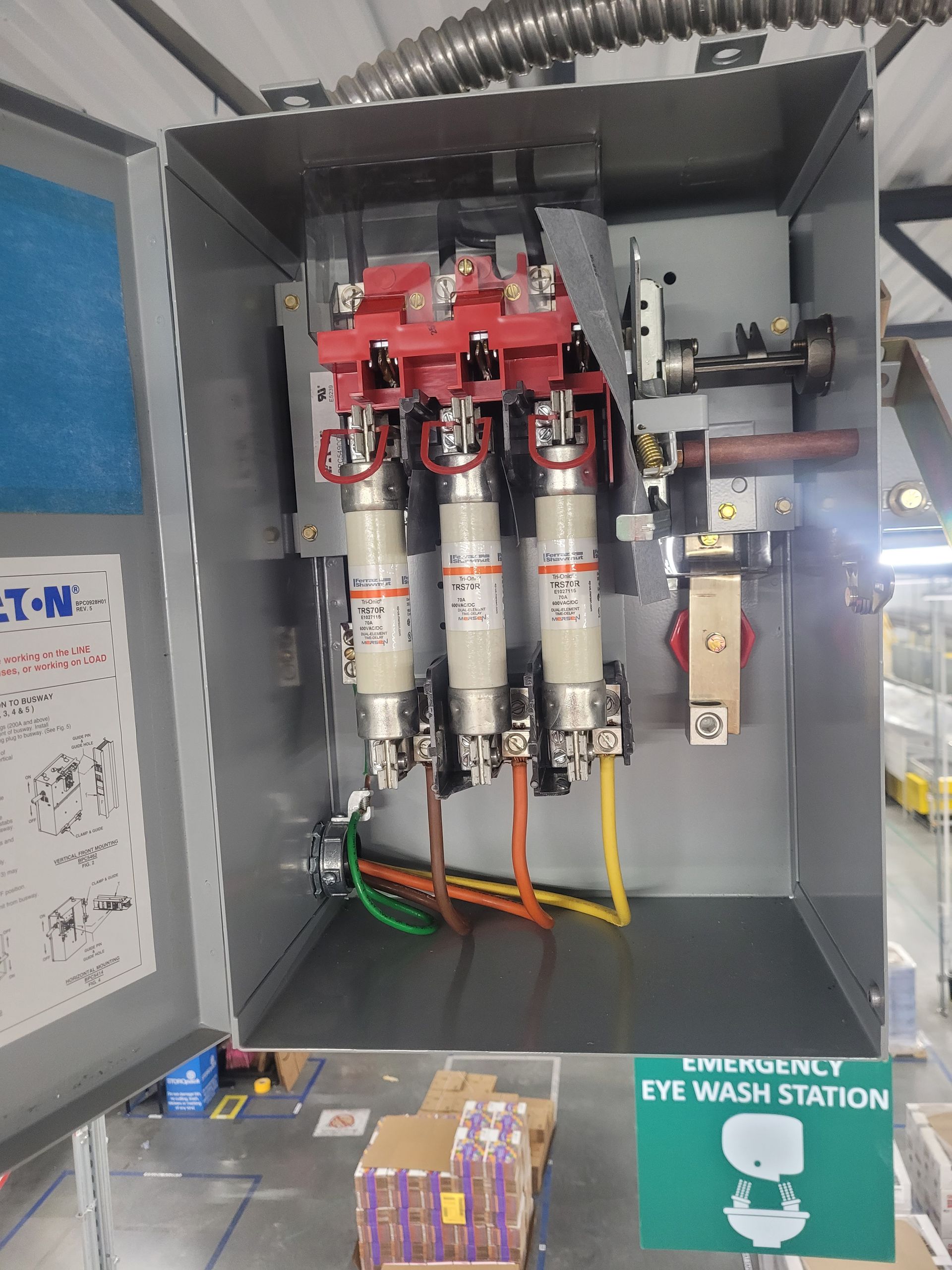 Open electrical fuse panel with three red switch fuses and labeled wiring, beside an eyewash station sign.