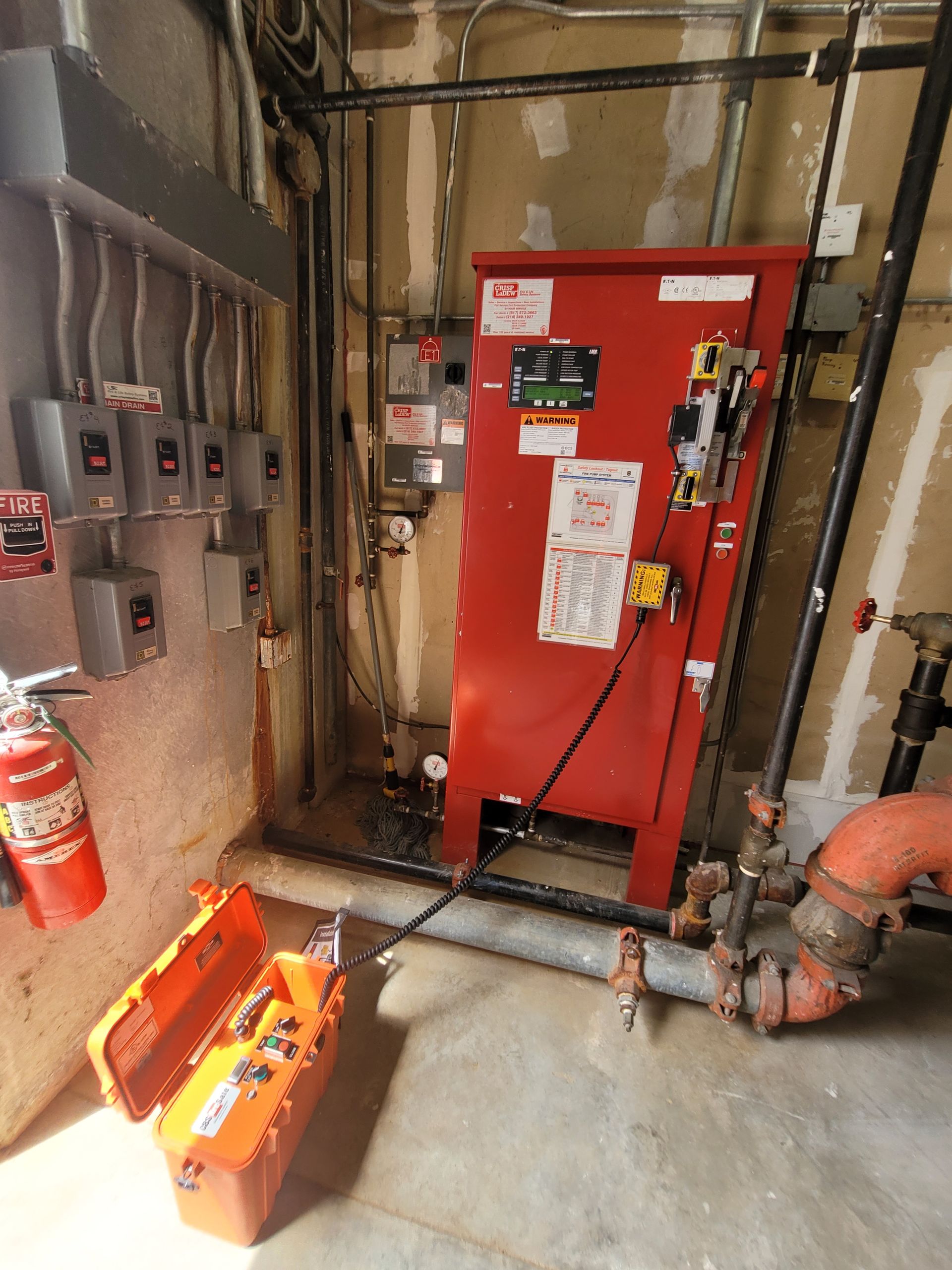 Industrial room with a red electrical control cabinet, wall panels, pipes, and a fire extinguisher on the floor.