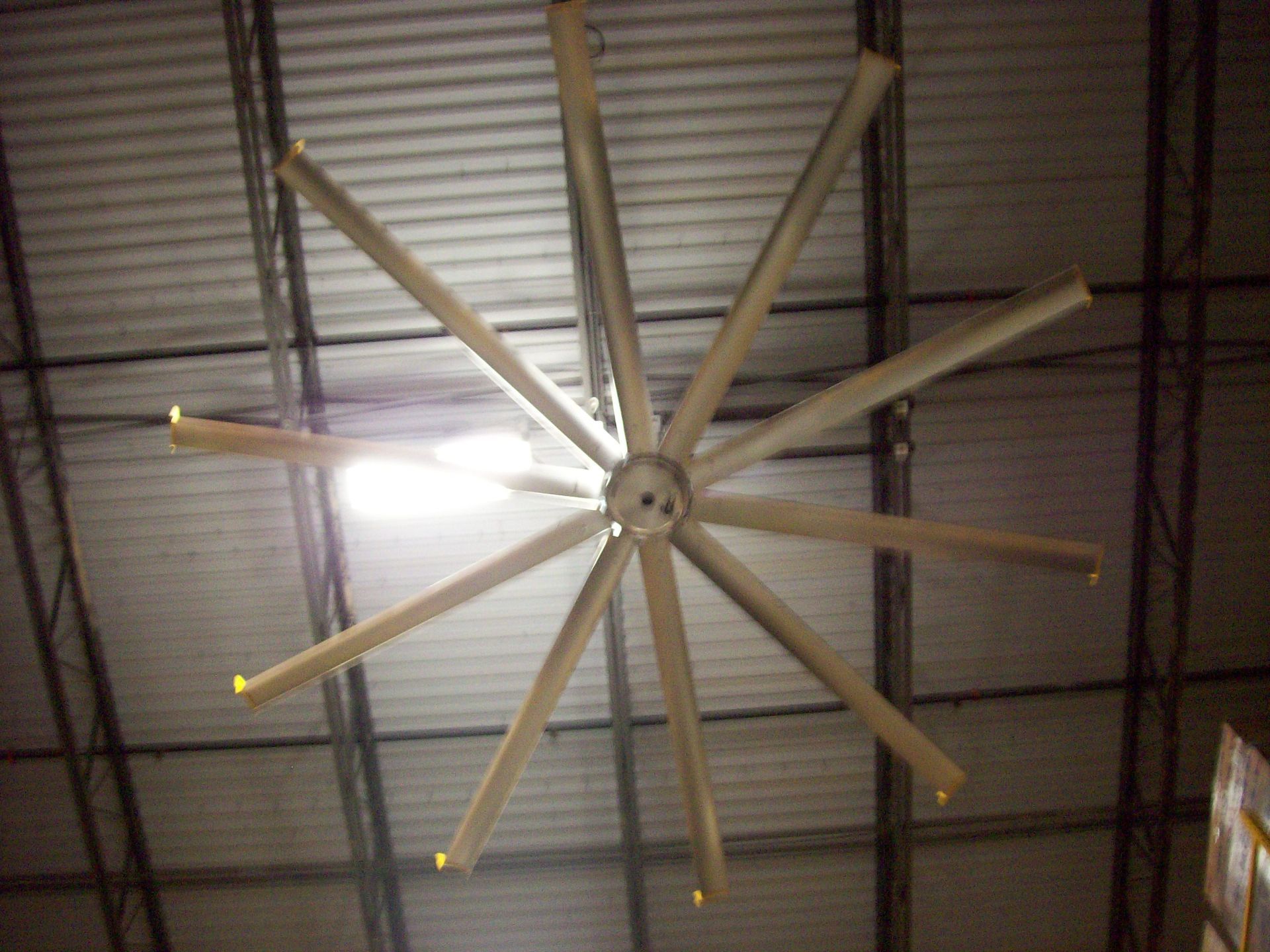 Ceiling fan with multiple blades mounted on a corrugated metal ceiling in a warehouse-like space