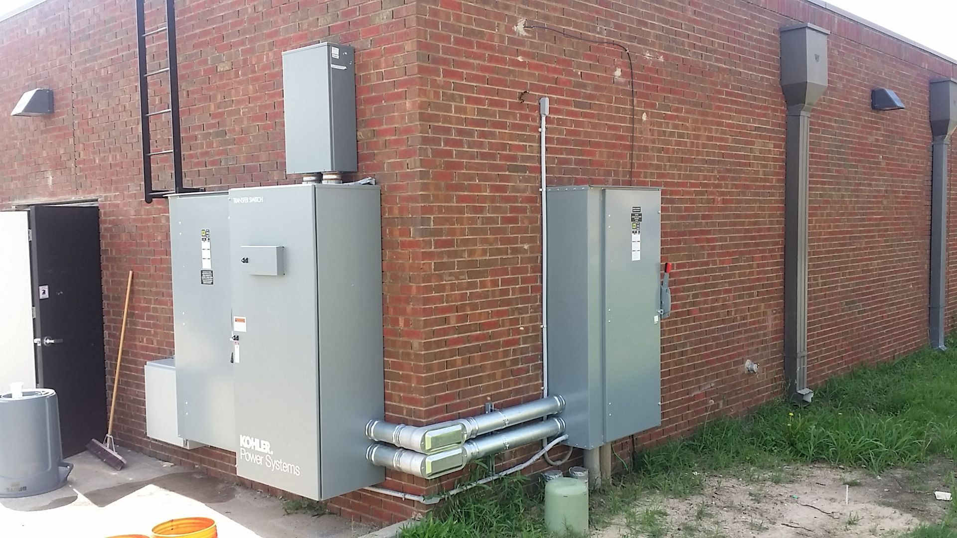 Gray utility/electrical boxes and pipes mounted on a brick building beside a grassy strip