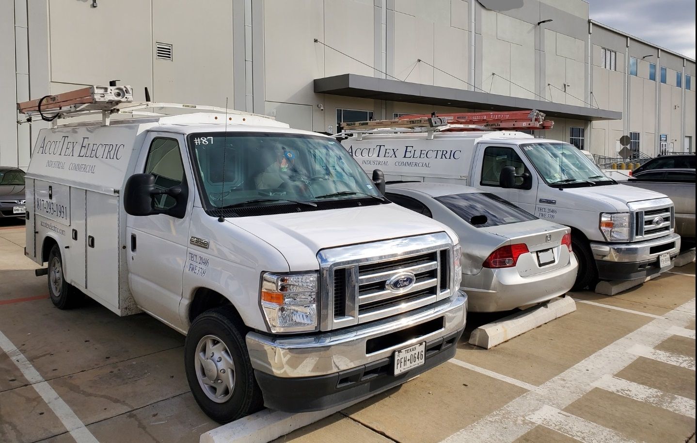 White utility vans parked beside a silver sedan in a lot outside a commercial building.