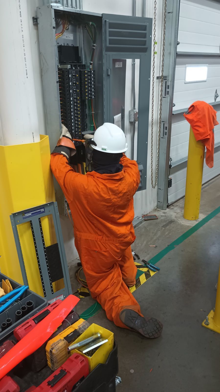 Worker in orange coveralls and white hard hat wiring an industrial electrical panel in a factory.