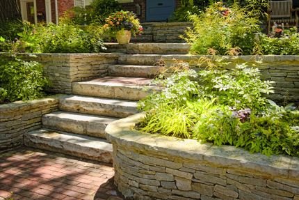 Stone garden steps surrounded by lush green plants and flower beds in sunlight