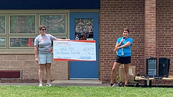 Two women are standing in front of a brick building holding a large check for Williamsport Elementary School donation.