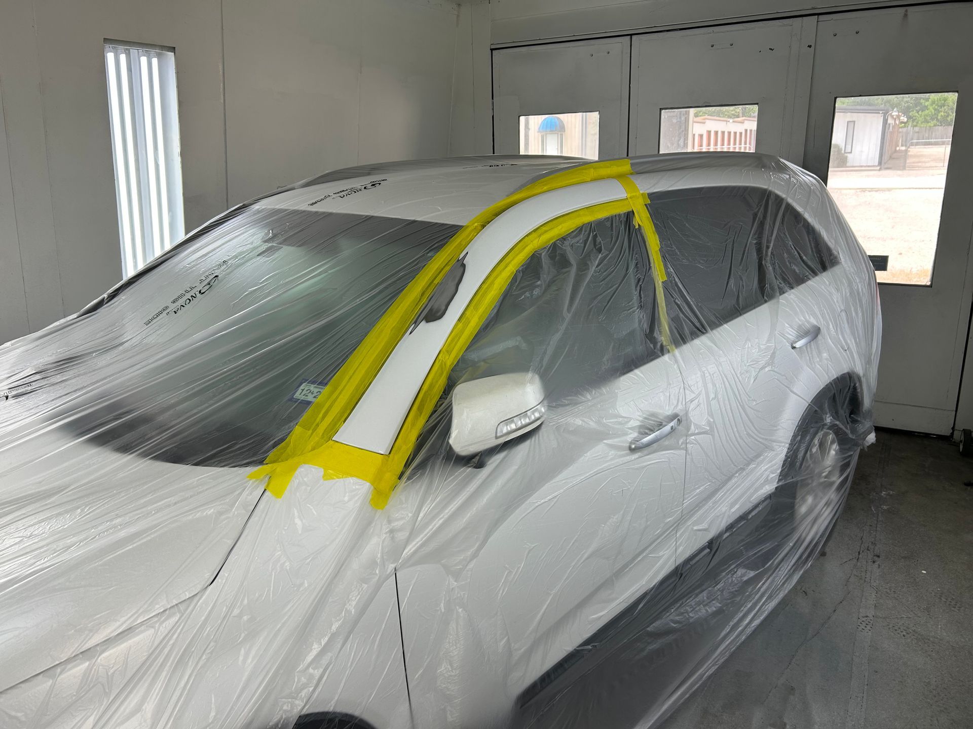 White car covered in plastic in a paint booth, taped trim ready for painting.