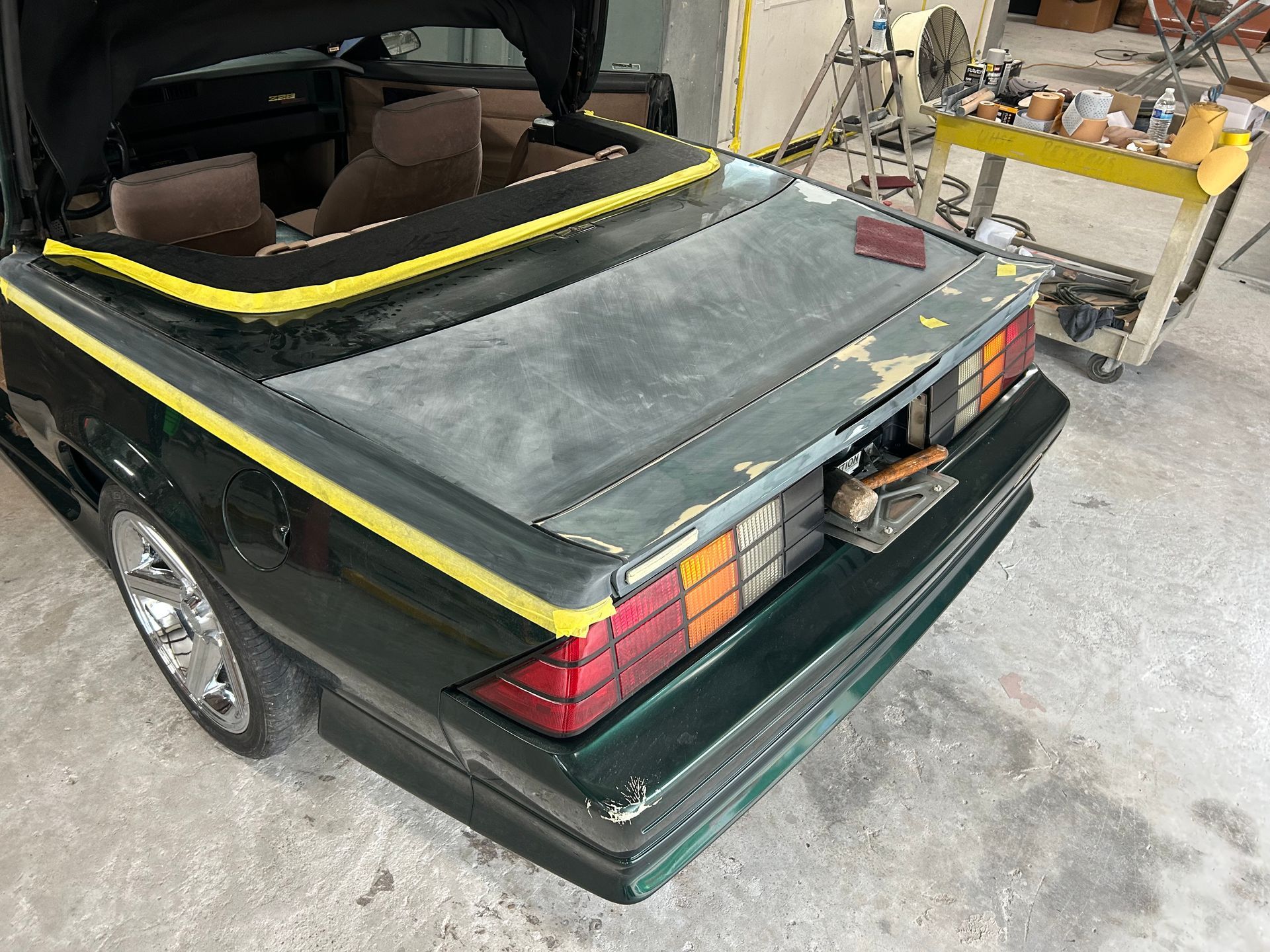 Green convertible car's rear end in a repair shop. Tail lights, faded paint, and a yellow stripe are visible.