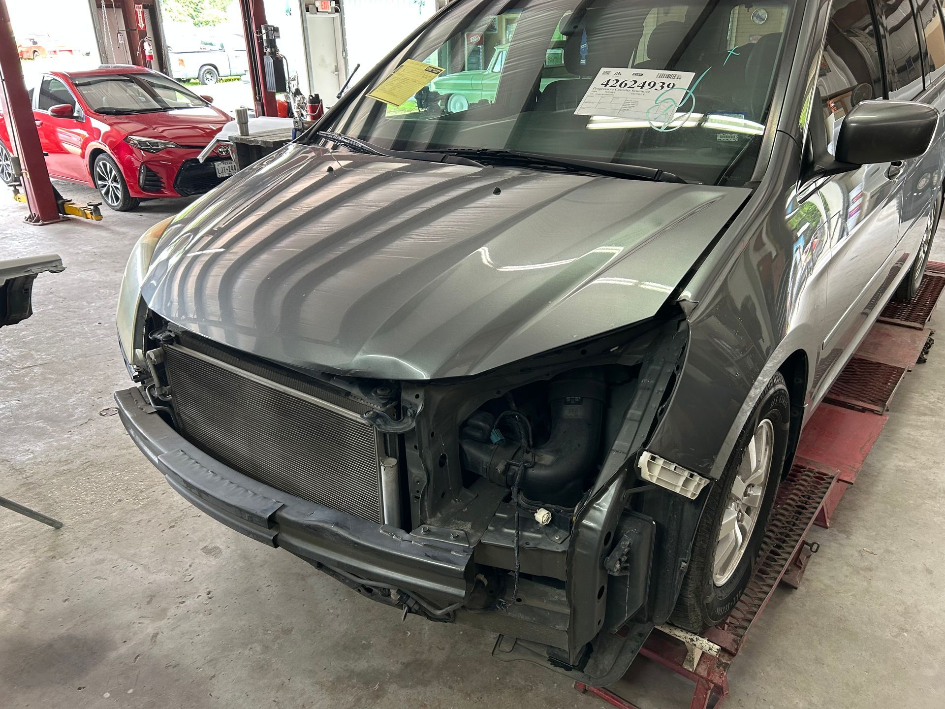 Gray minivan with front-end damage in an auto repair shop, next to a red car.