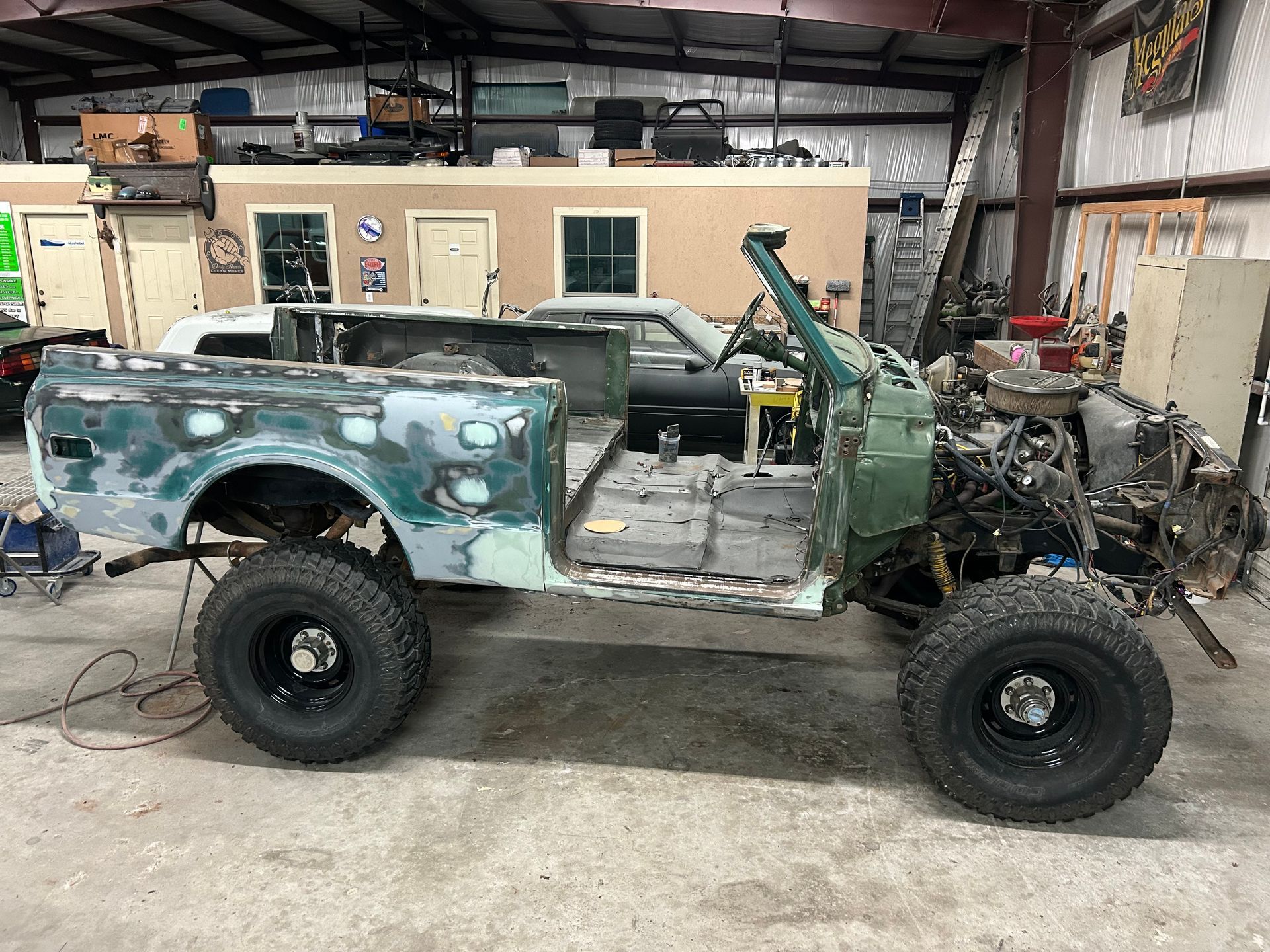 Green and grey truck undergoing restoration in a workshop, missing doors, with large tires.