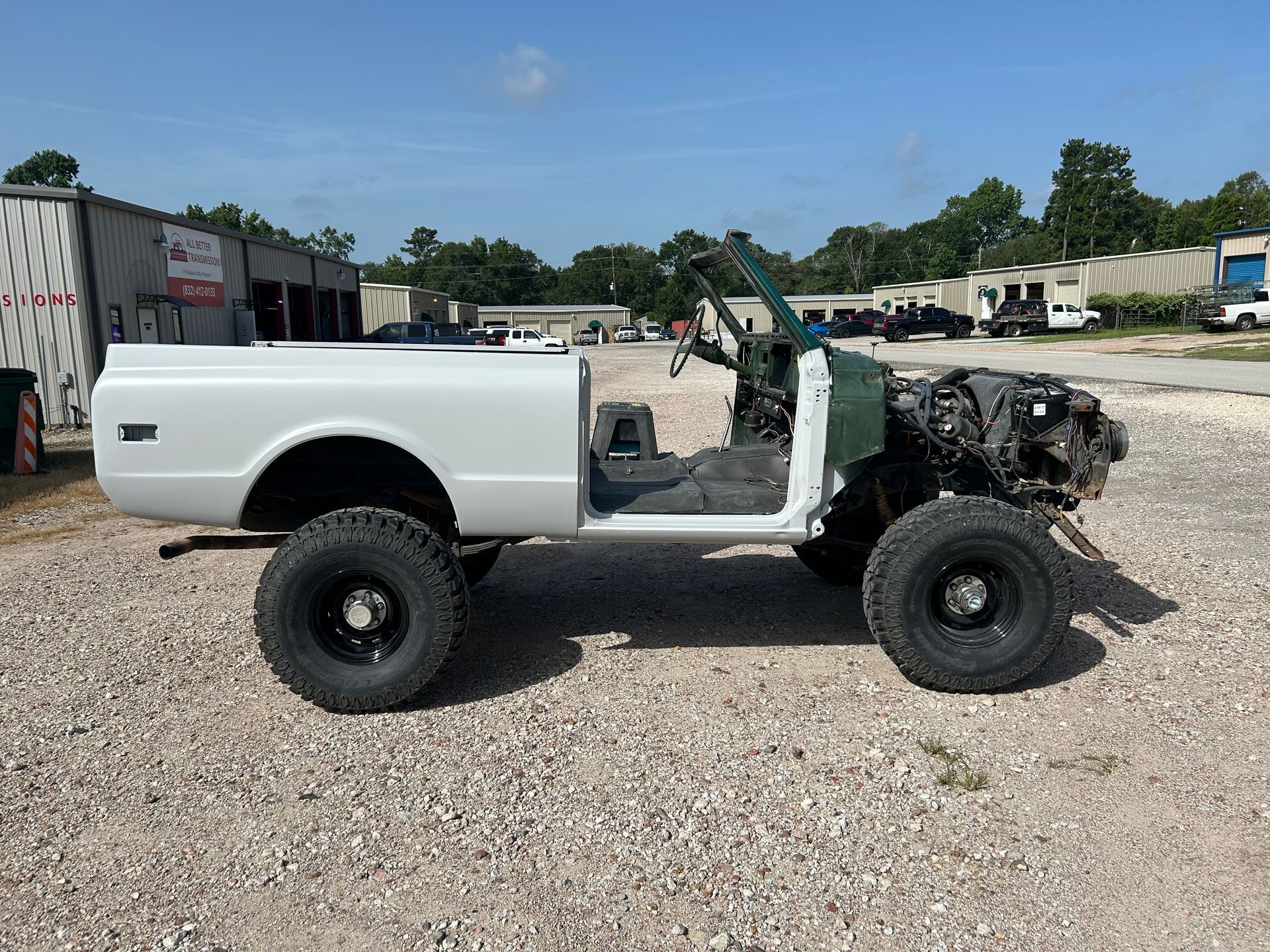 White pickup truck being rebuilt, sits on large tires, with green cab, in a gravel lot.