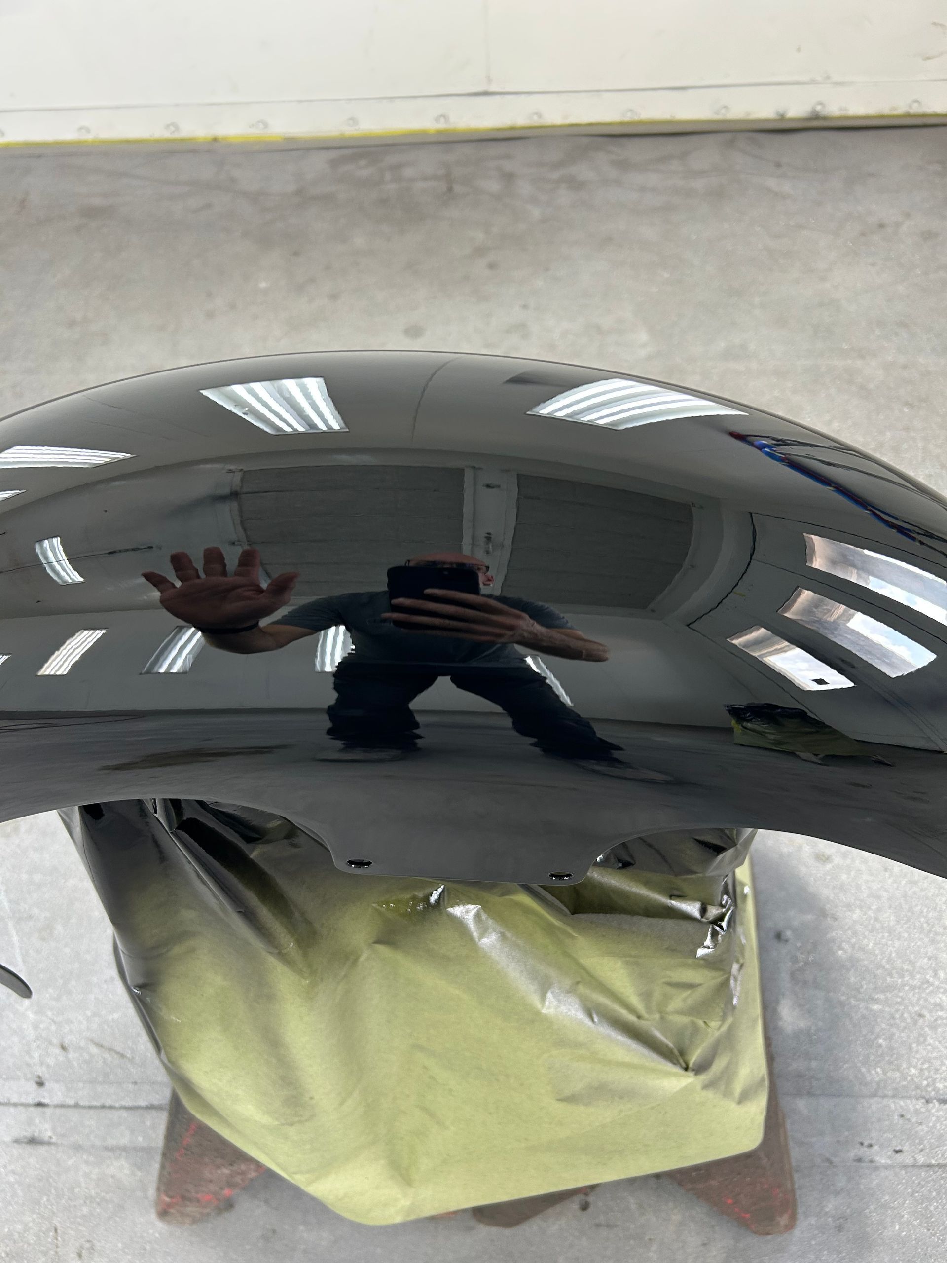 Black car fender reflecting a person in a workshop, holding up a hand.