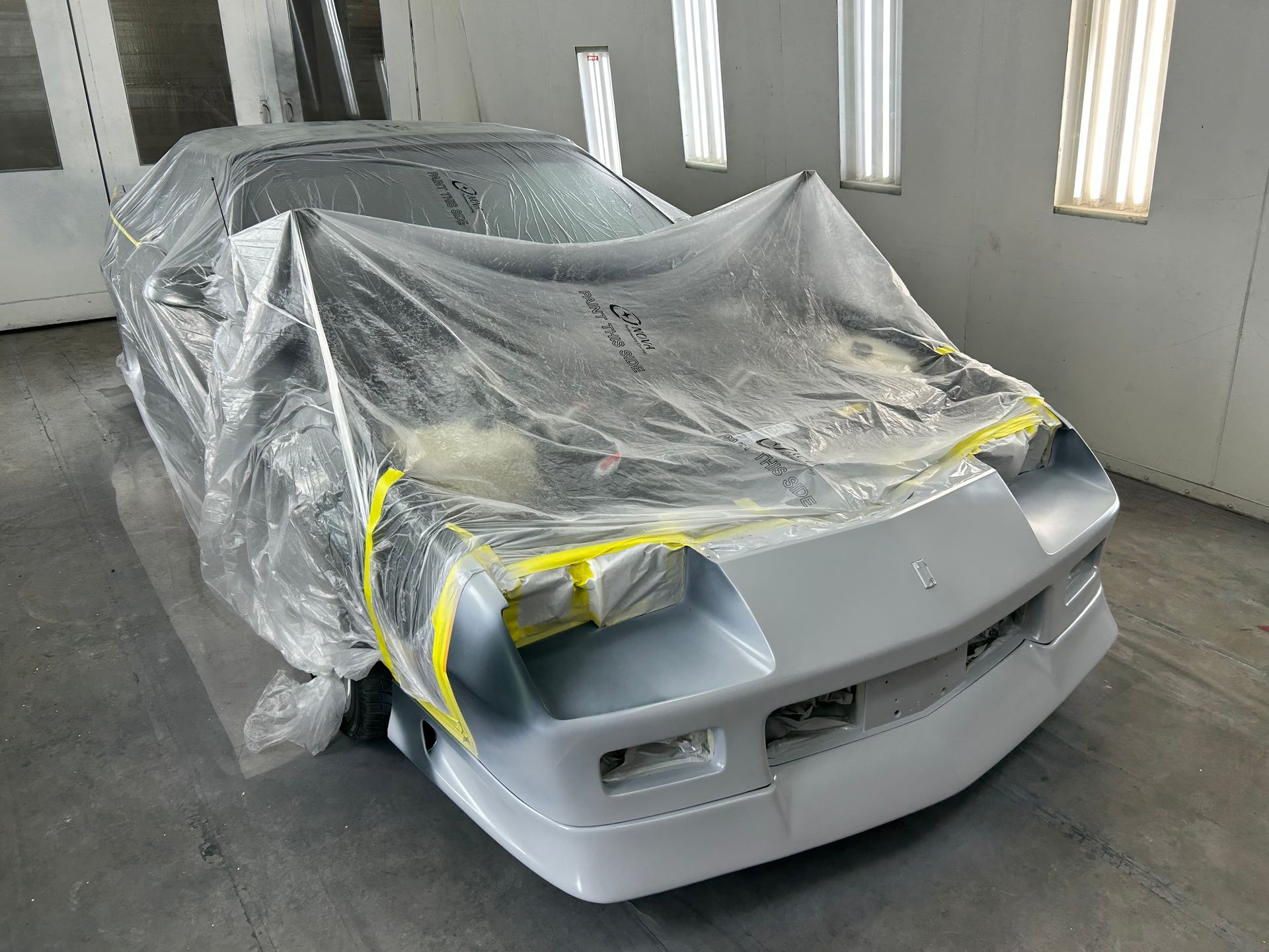 Gray sports car in a paint booth, covered in plastic and taped off, ready for painting.