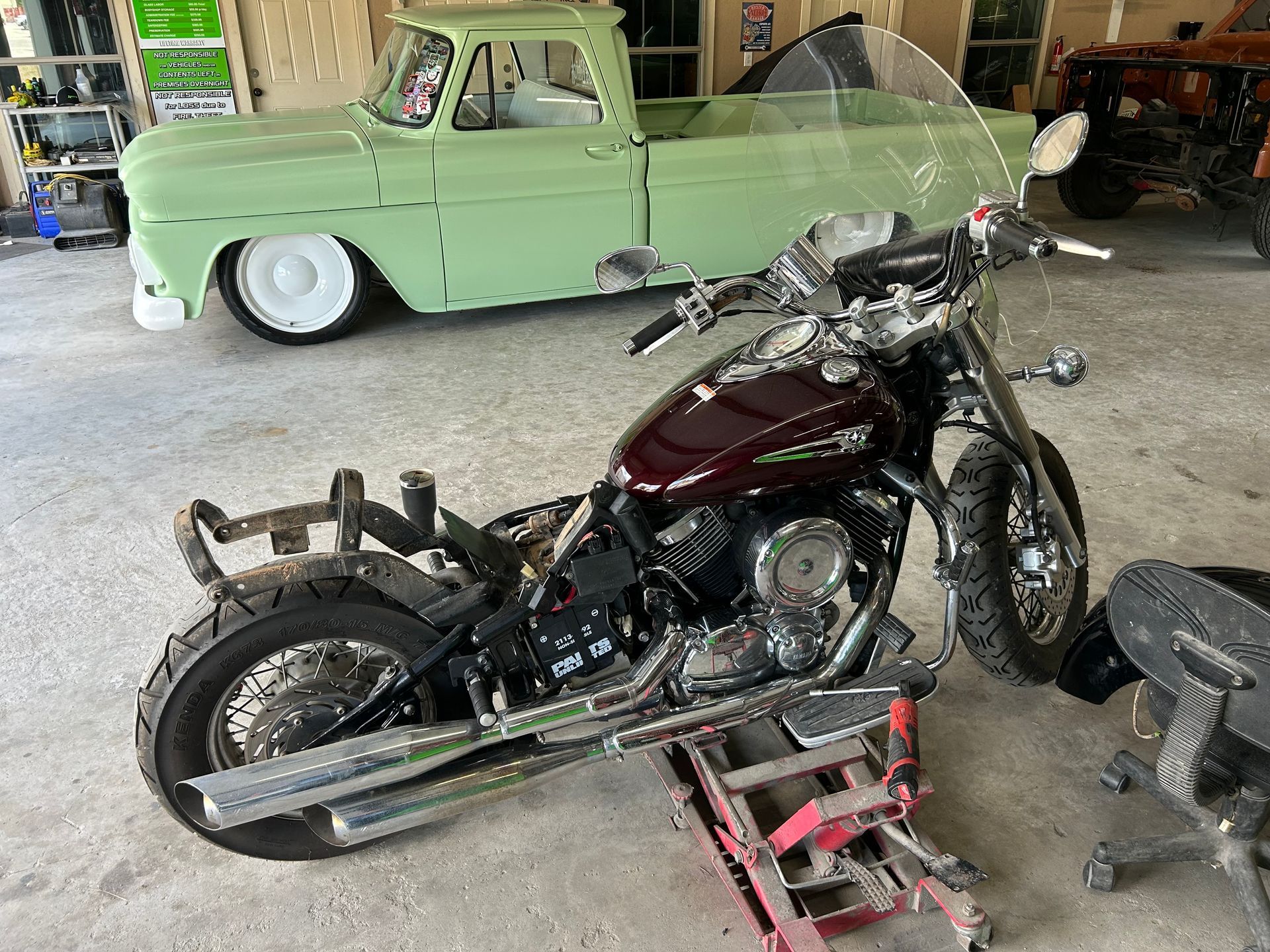 Maroon motorcycle on a lift in a garage; a green classic pickup truck is in the background.