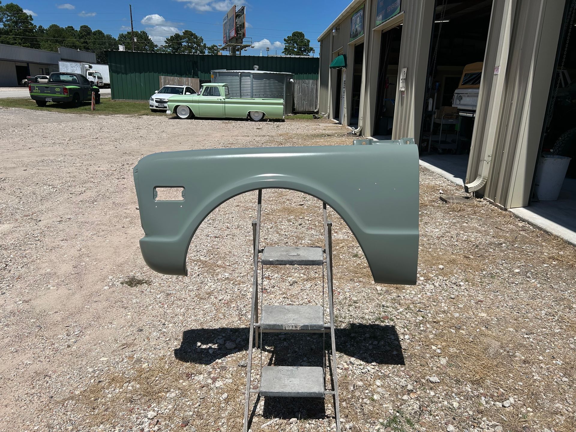 A grey truck fender rests above a step ladder on gravel; other trucks and a building are in the background.