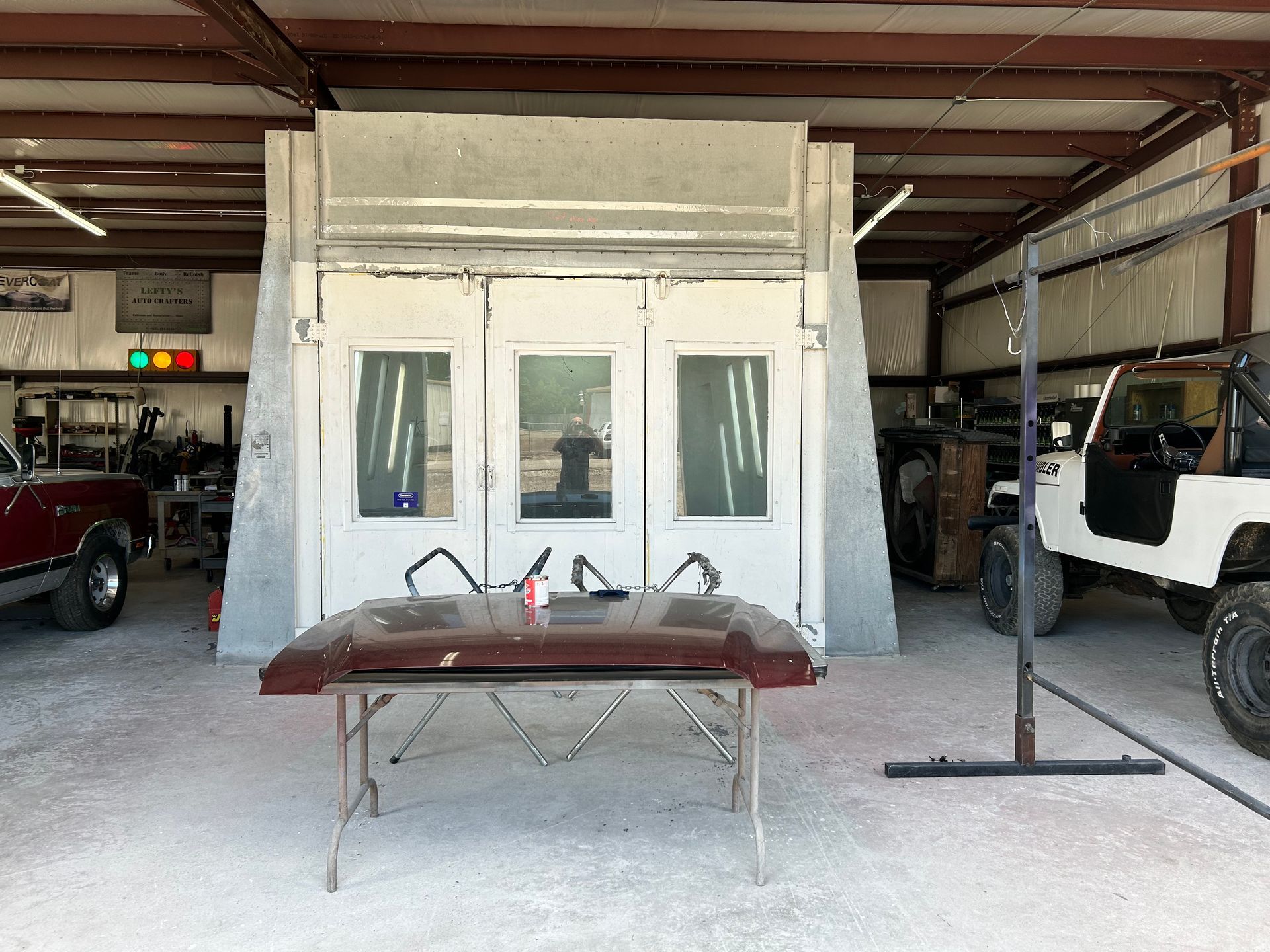 A car hood on a table in front of a paint booth in a garage; a Jeep and truck are visible.