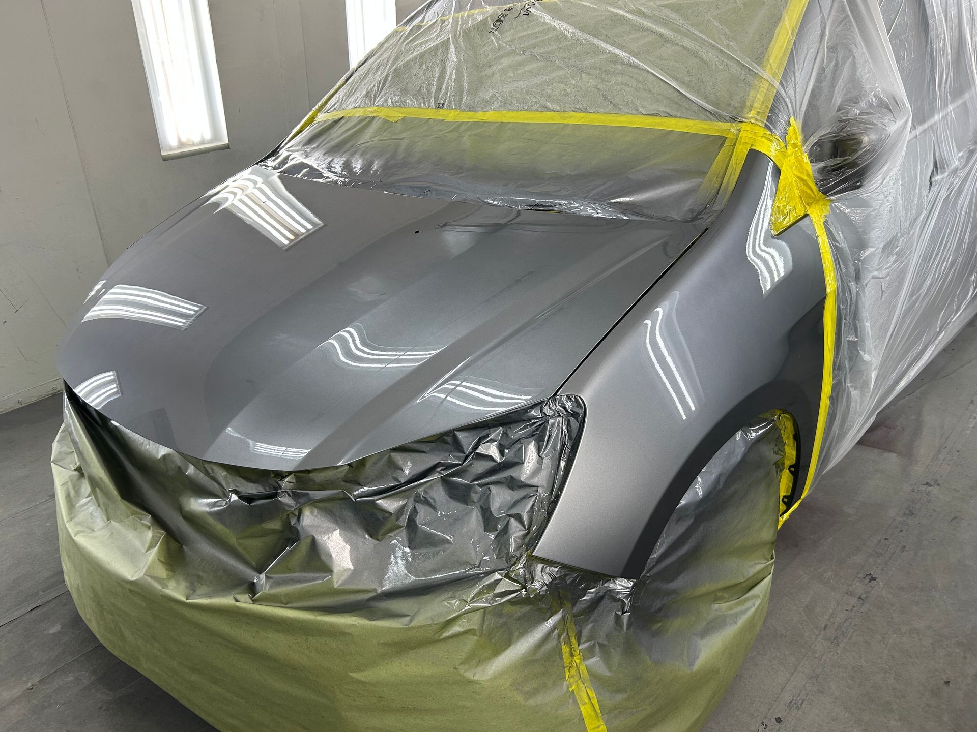 A gray car in a paint booth, masked with plastic and yellow tape, ready for paint.