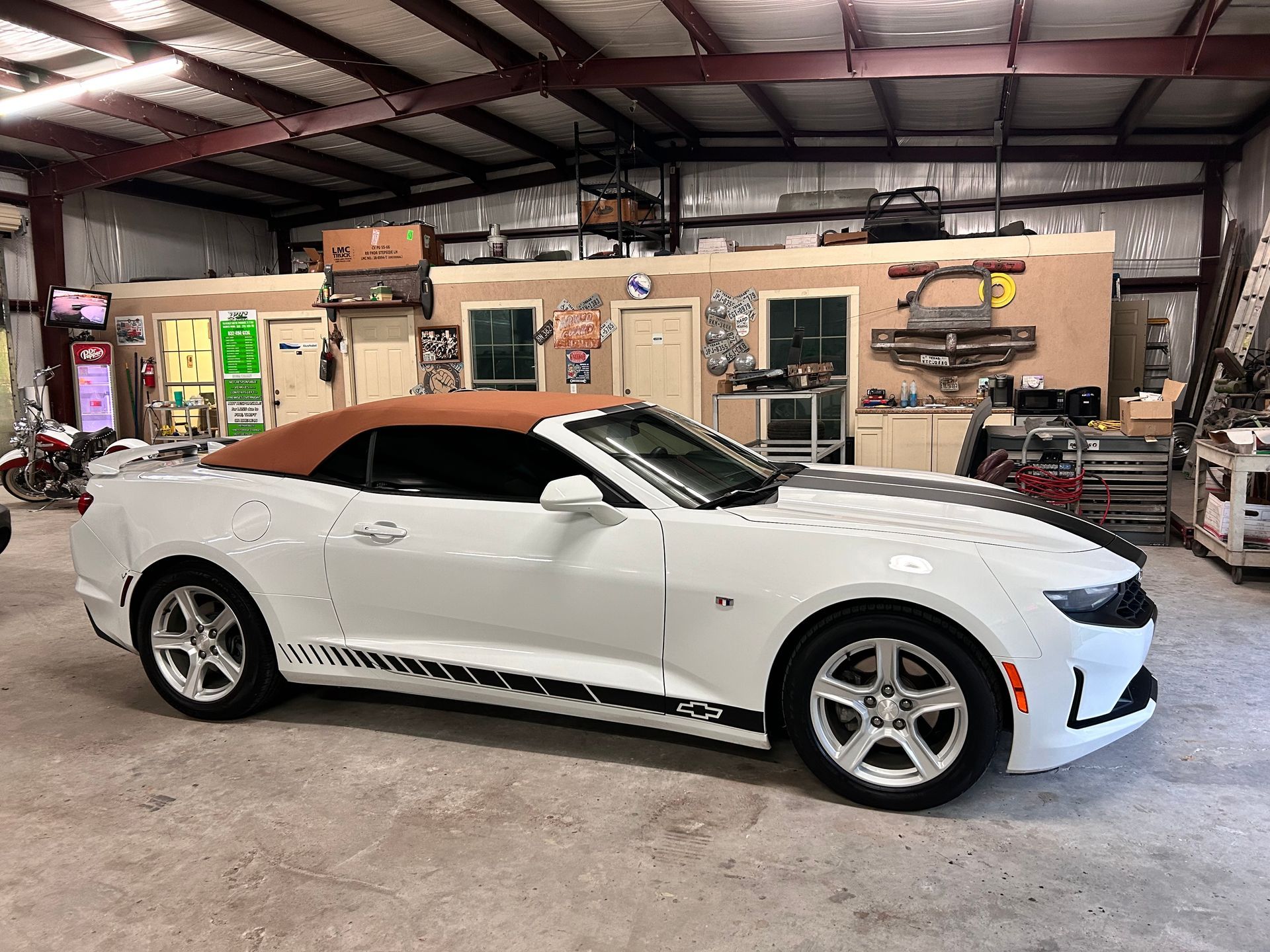 White convertible Camaro with brown top, black racing stripes, parked inside a garage.