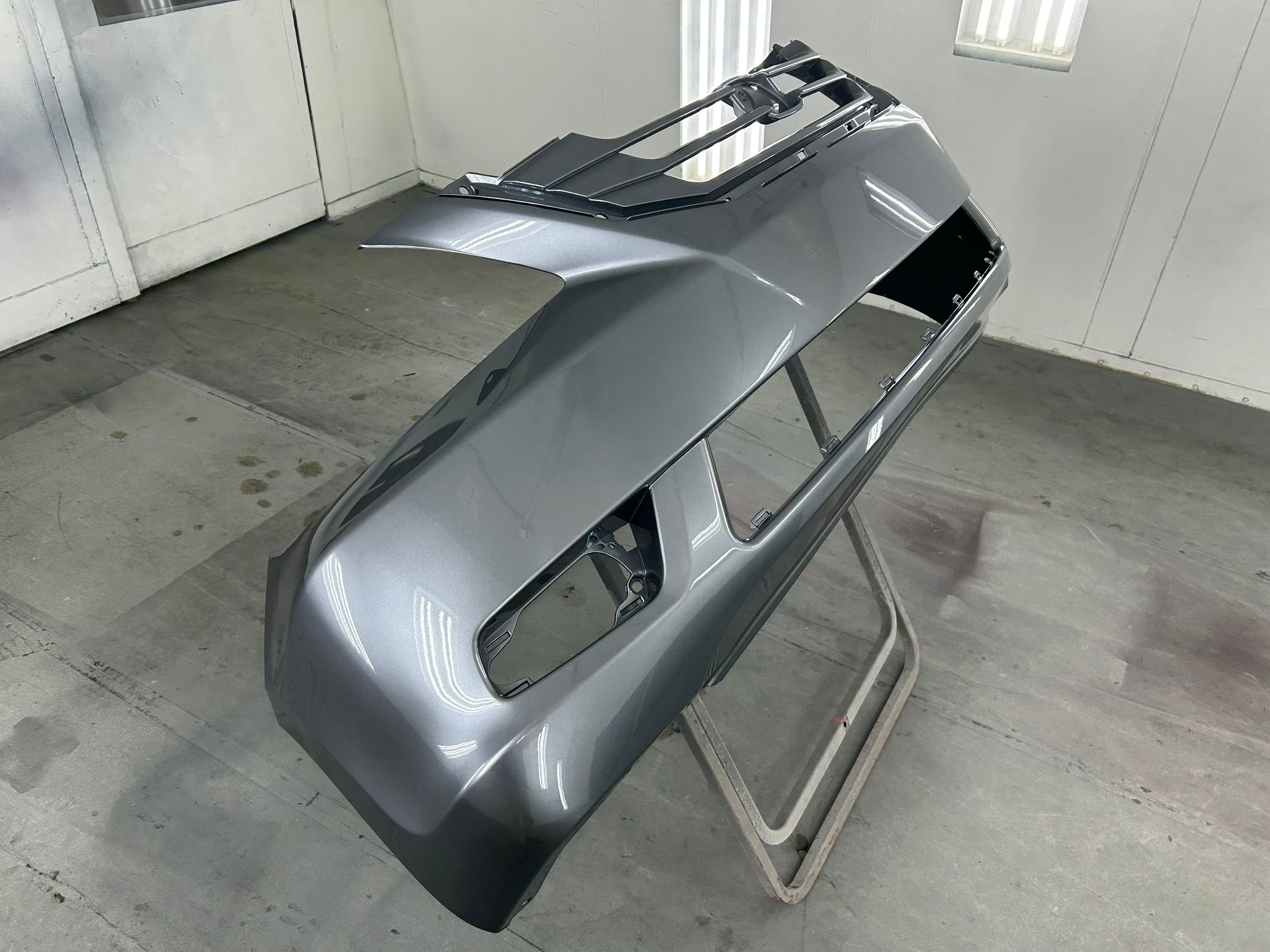 Gray car bumper, freshly painted, sitting on a stand in a spray booth.