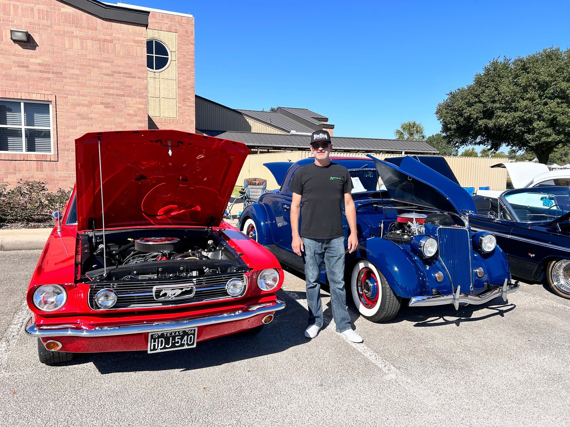 Man stands between a red Mustang and a blue classic car, all with open hoods, outdoors.