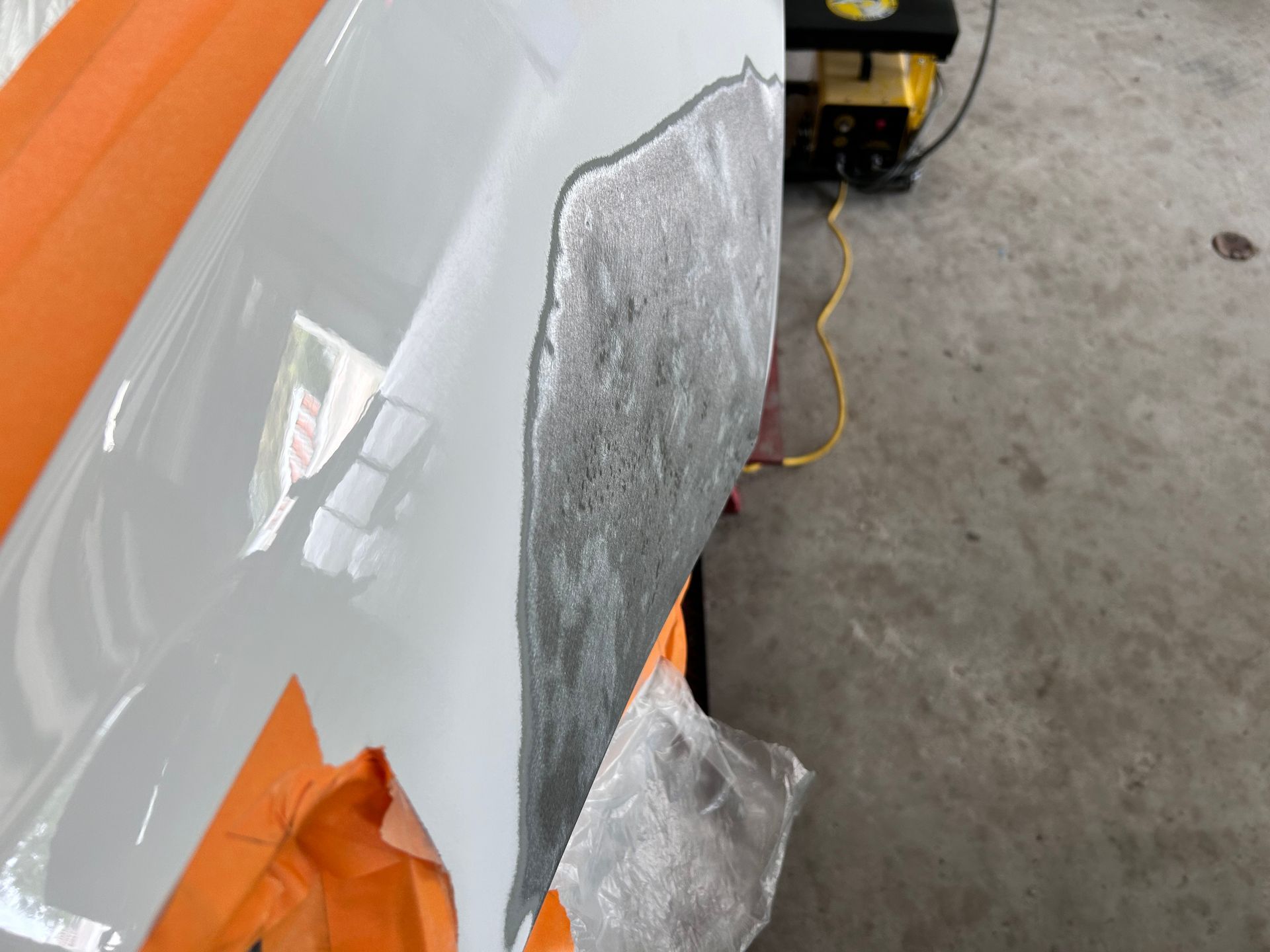 White car panel with gray primer patch, orange tape, and a mechanical pump.