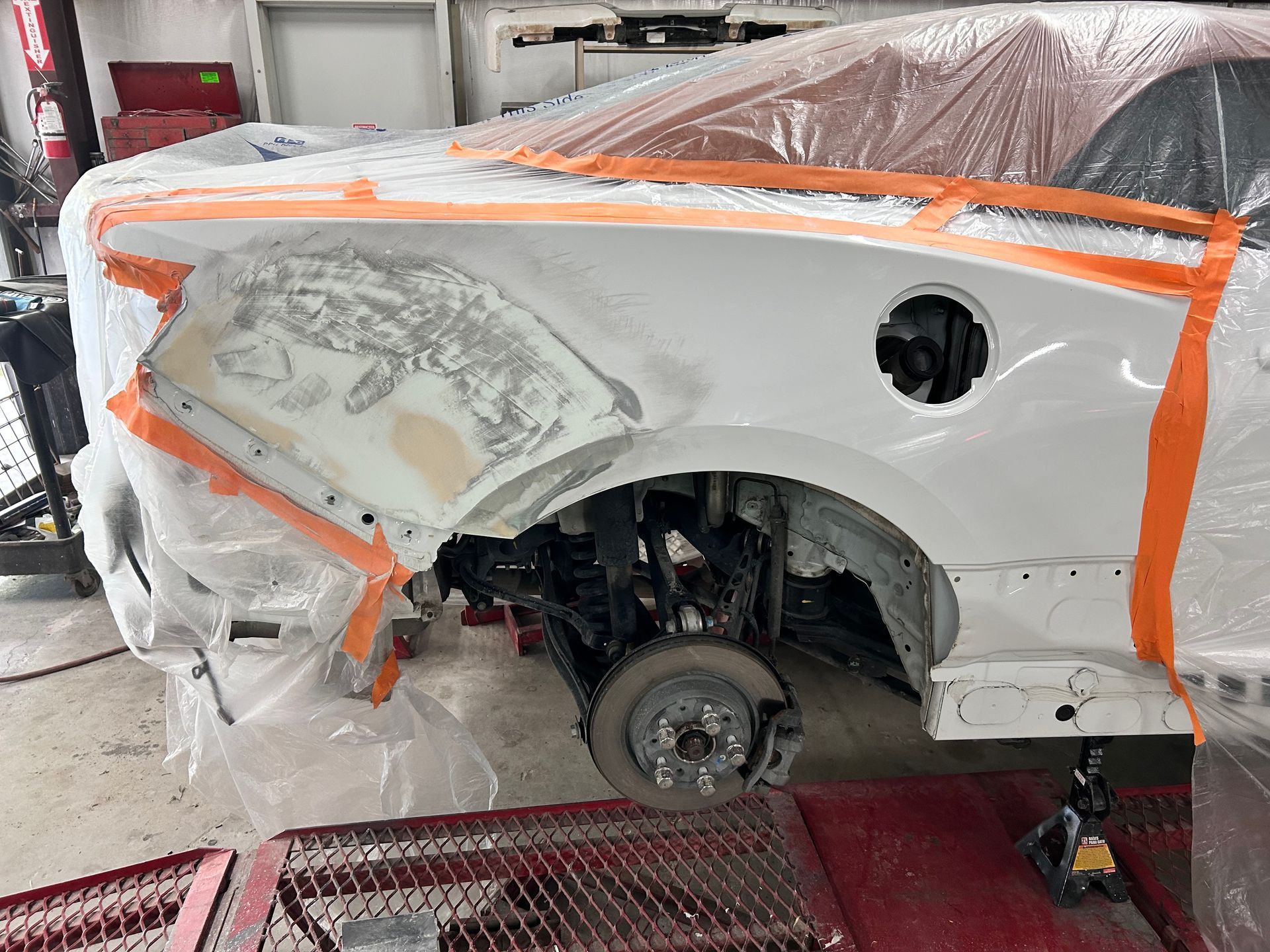 White car fender being repaired, taped, and covered in plastic in a garage setting.